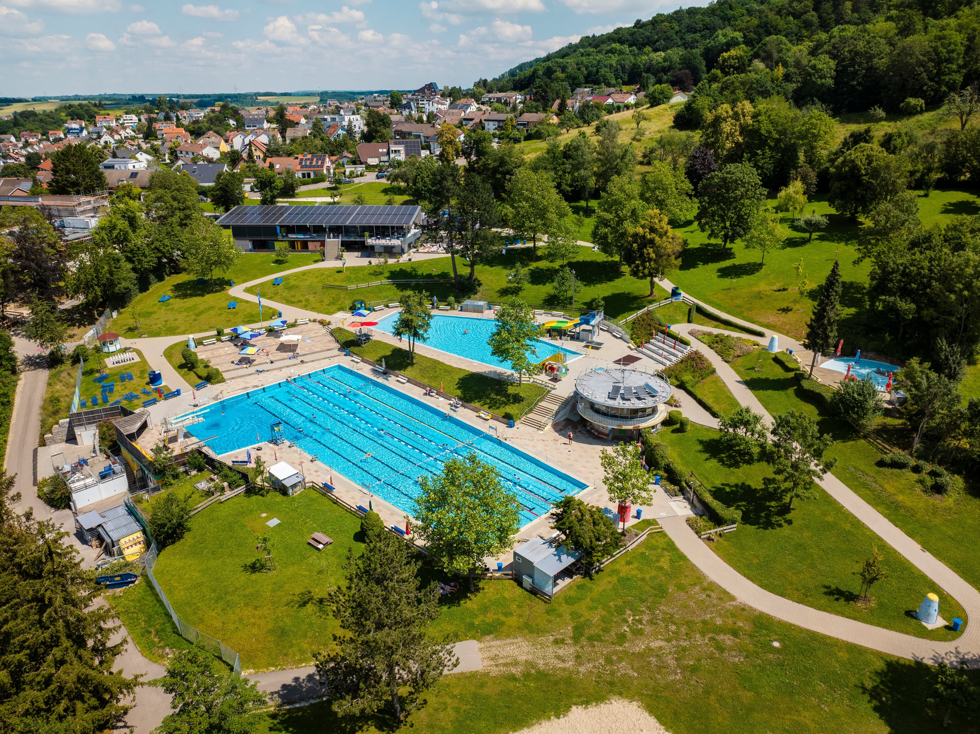 Eine Luftaufnahme des Spieselfreibads mit den drei Schwimmbecken, Liegewiese und Bäumen. Im Hintergrund ist der Teilort Wasseralfingen zu sehen sowie