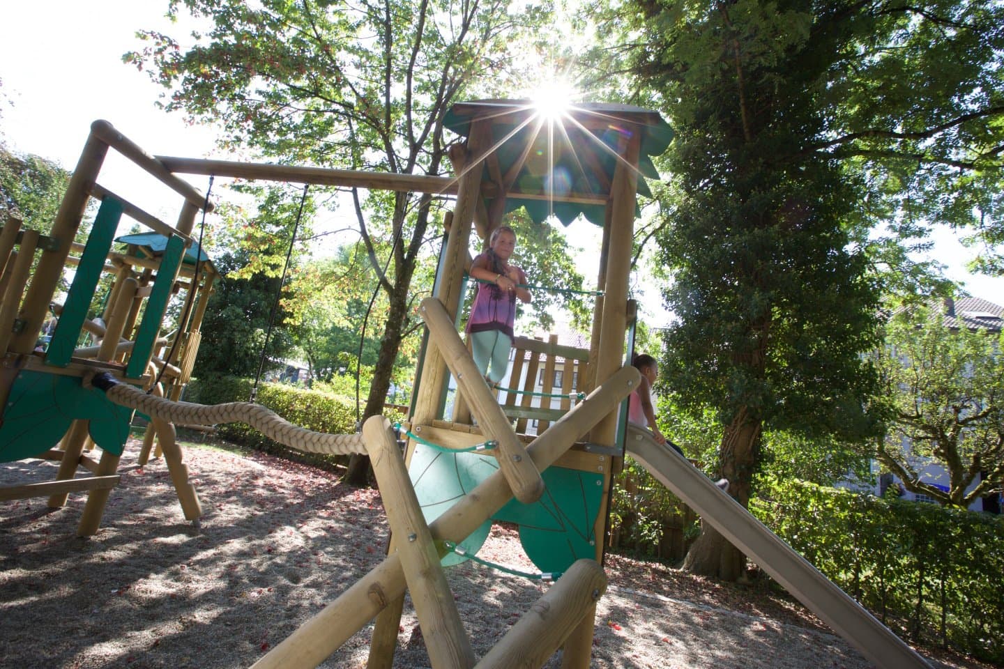 ein Kletterturm mit Rutsche und einer kleinen Hängebrücke zu einem weiteren kleineren Turm aus Holz gebaut, mit Gegenlicht fotografiert, im Anna-Kym Garten