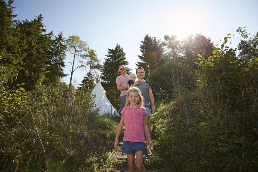 Das Bild zeigt eine Familie beim Wandern zwischen Wiesen und Sträuchern. Im Hintergrund steht ein weißes Tipi am Waldrand.