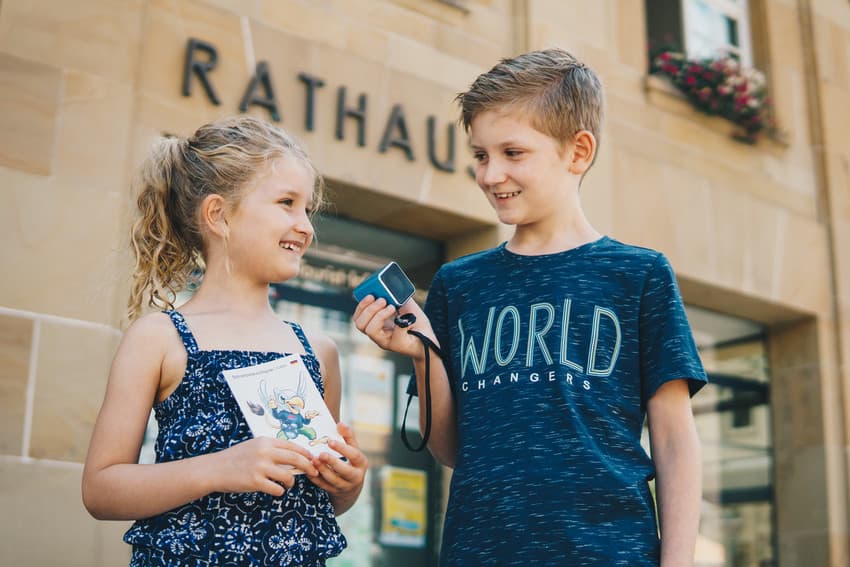 Auf dem Bild sind zwei Kinder vor dem Rathaus Schramberg zu stehen. Die Kinder halten das Audioguide-Tourenprospekt wie die Lautsprecher in der Hand.