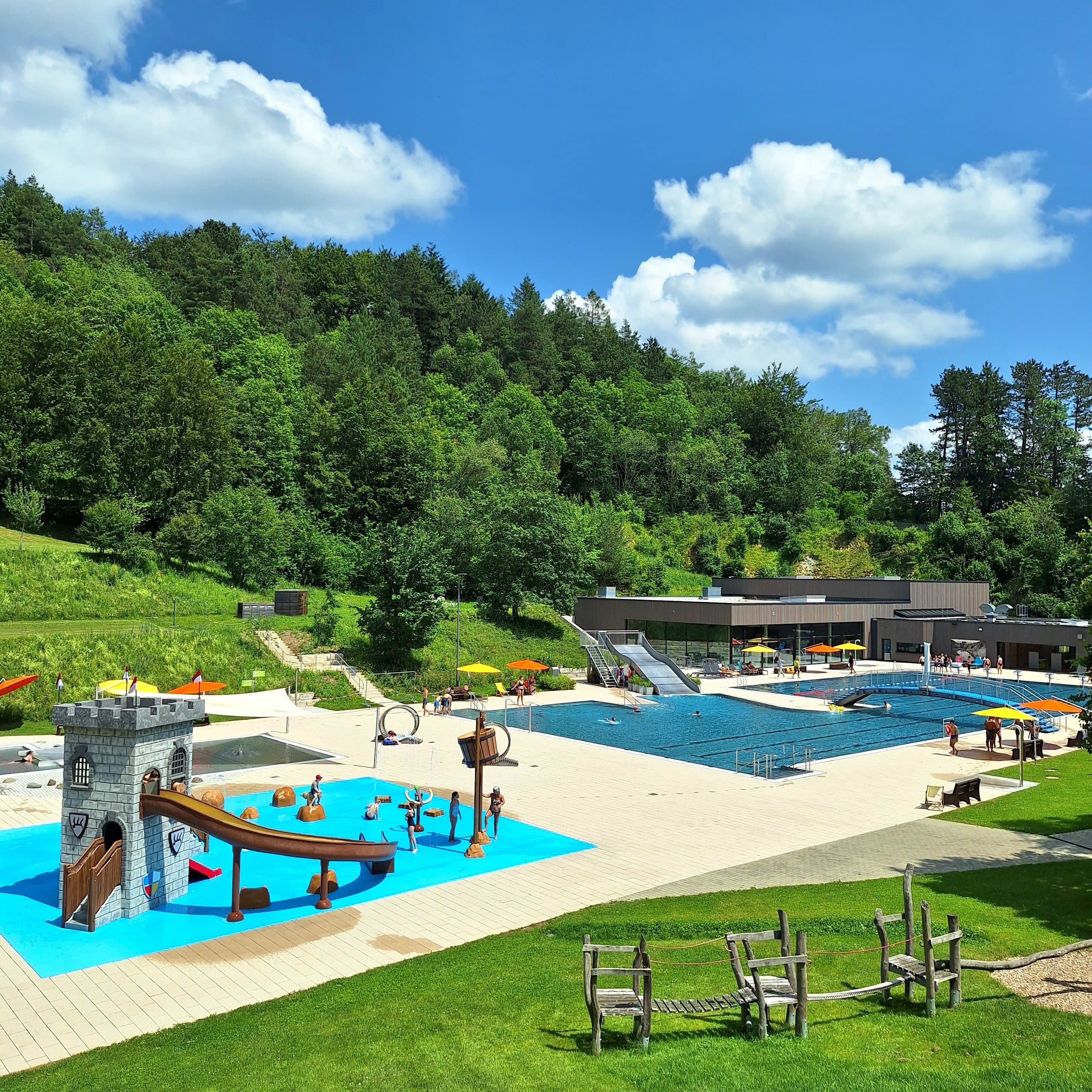 Ein Freibad mit einem großen Becken, einer Wasserrutsche, einem kleinen Kinderbecken, einem Wasserspielplatz, vielen Liegewiesen, einem Kiosk und weiteren Spielmöglichkeiten. Überall tummeln sich Leute. Im Hintergrund sind Bäume.