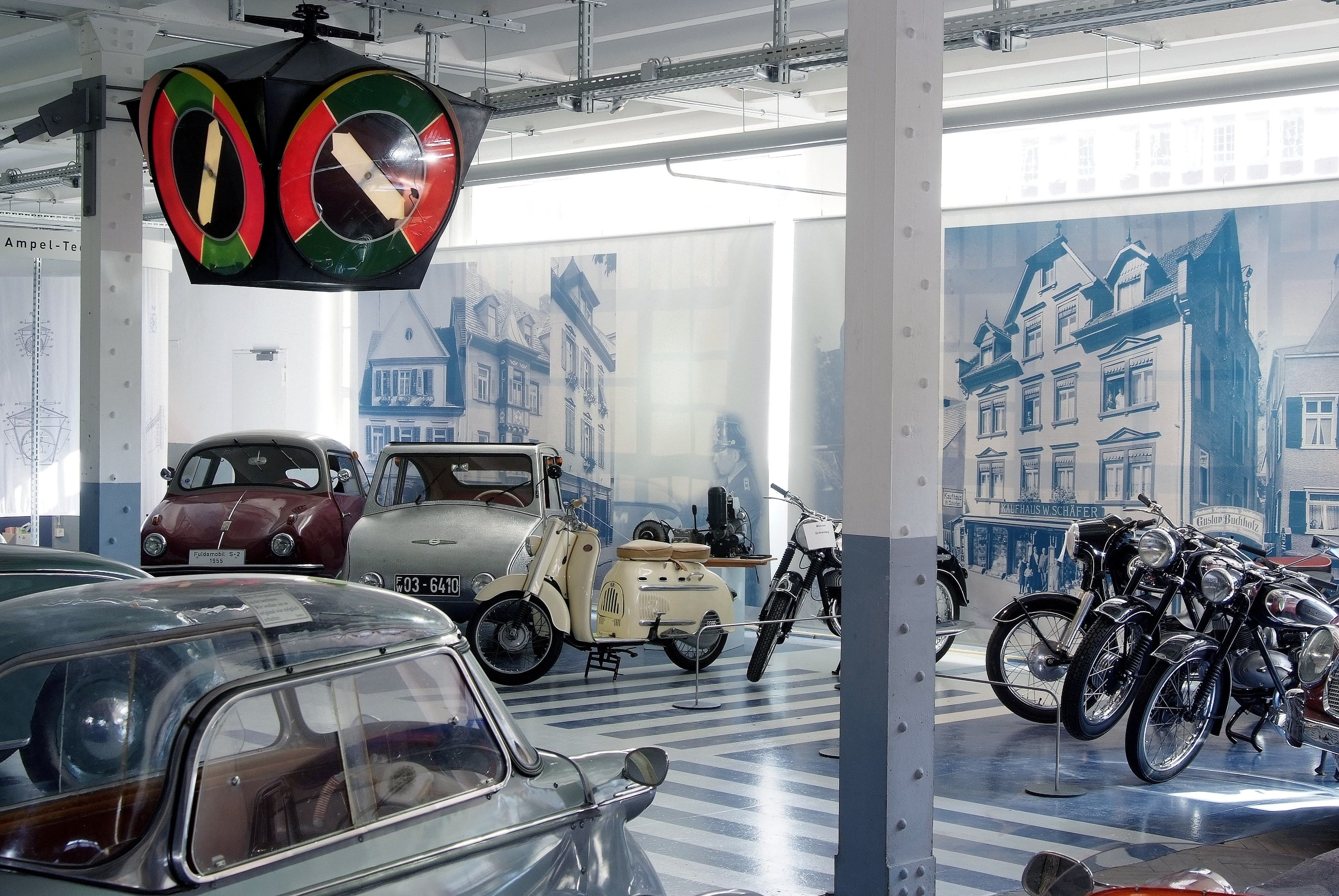 Auf dem Bild ist ein Ausstellungsraum mit historischen Fahrzeugen und Motorrädern, darunter ein VW Käfer und eine Vespa, zu sehen.