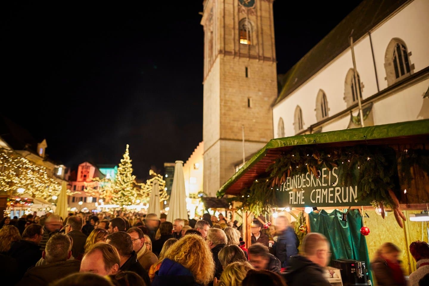 Ein beleuchteter Weihnachtsmarkt mit vielen Besuchern und einem Verkaufsstand direkt vor einer Kirche.