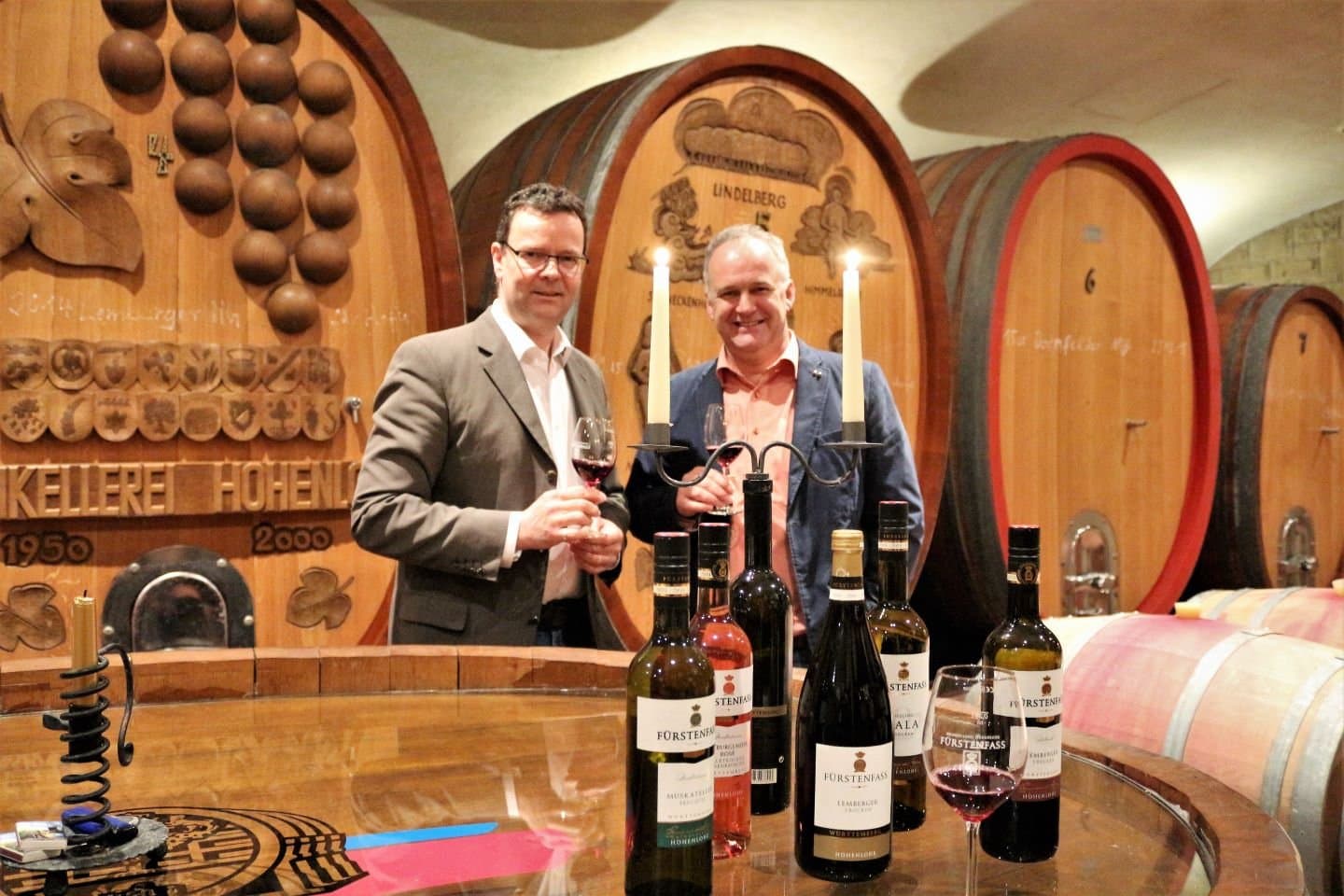 Zwei Männer halten ein Glas Wein in der Hand und stehen vor großen, schmucken Holzfässern