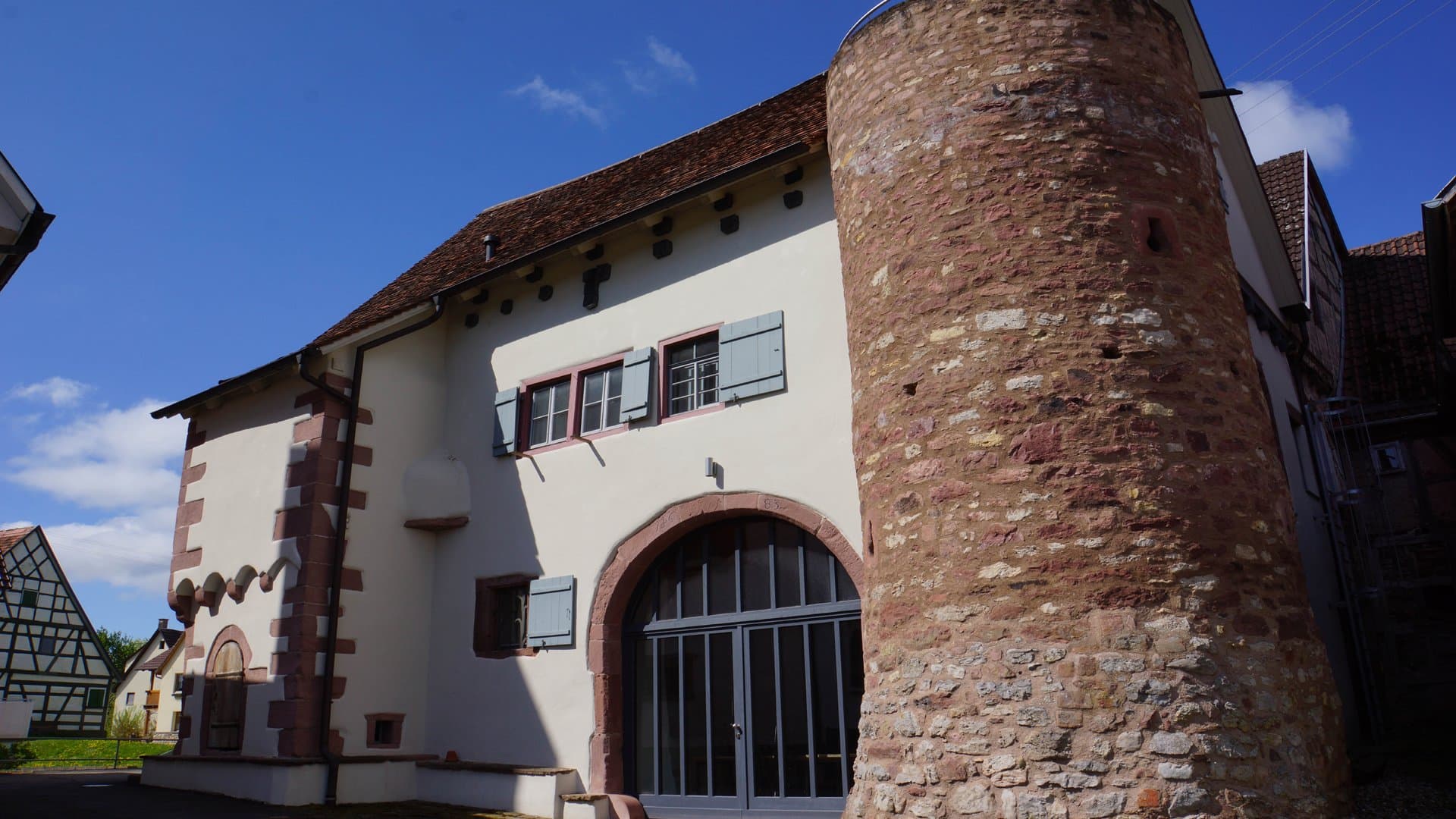 Mittelalterliches Gebäude mit rundem Turm, braunen Steinen und weißen Wänden, unter blauem Himmel.
