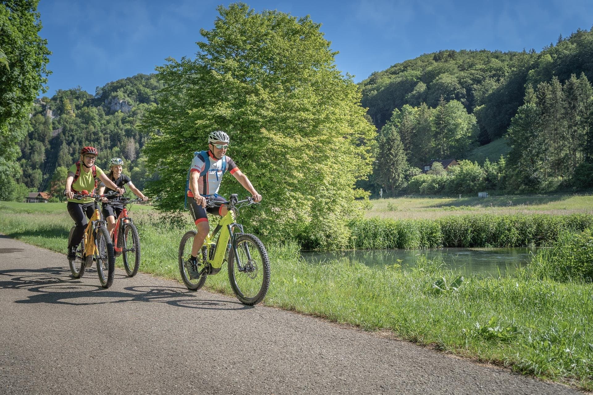 Die e-Bike Tour führt von Münsingen dursch Große Lautertal nach Wimsen und Marbach im Biosphärengebiet Schwäbische Alb. Drei Radfahrer*innen fahren mit E-Bikes auf einem Teerweg an einem Fluss entlang. Im Hintergrund ist Wald ud einige Felsen.