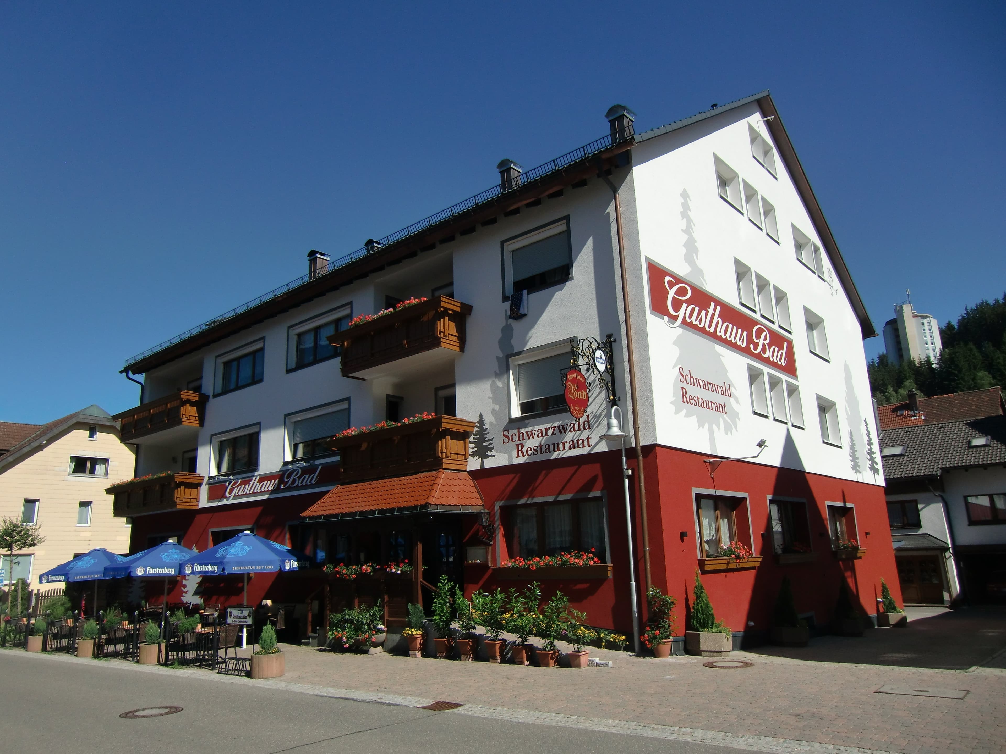Außenansicht des Gasthaus Bad mit Eingangsbereich und einer Terrasse mit Sonnenschirmen und Gartenmöbeln.