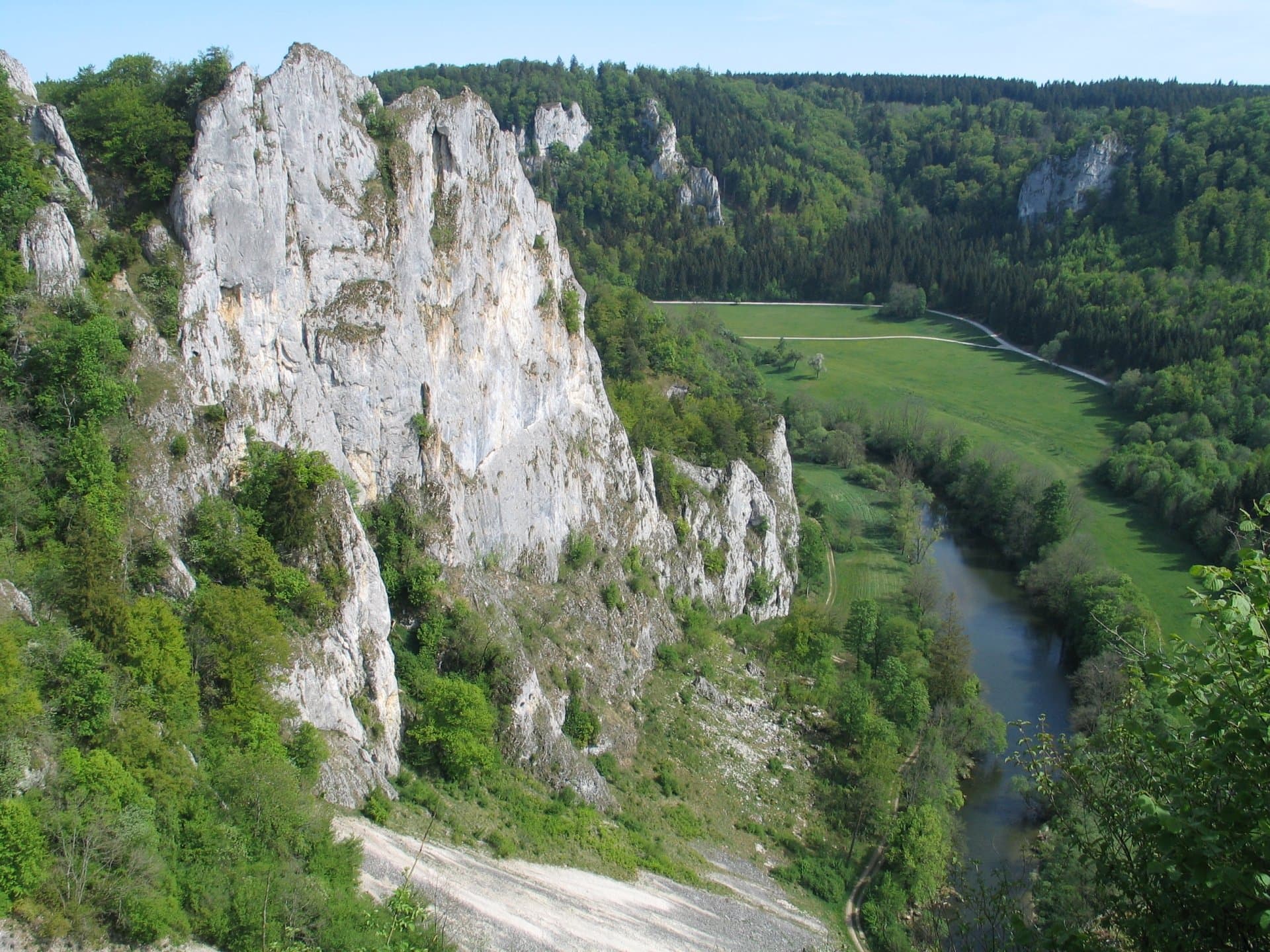 Großes Felsmassiv mit Büschen und Bäumen umgeben. Am Fuß des Felsens ein Flusstal. Rechts des Flusses eine Wiese, ein Weg und bewaldete Hänge mit Felsen