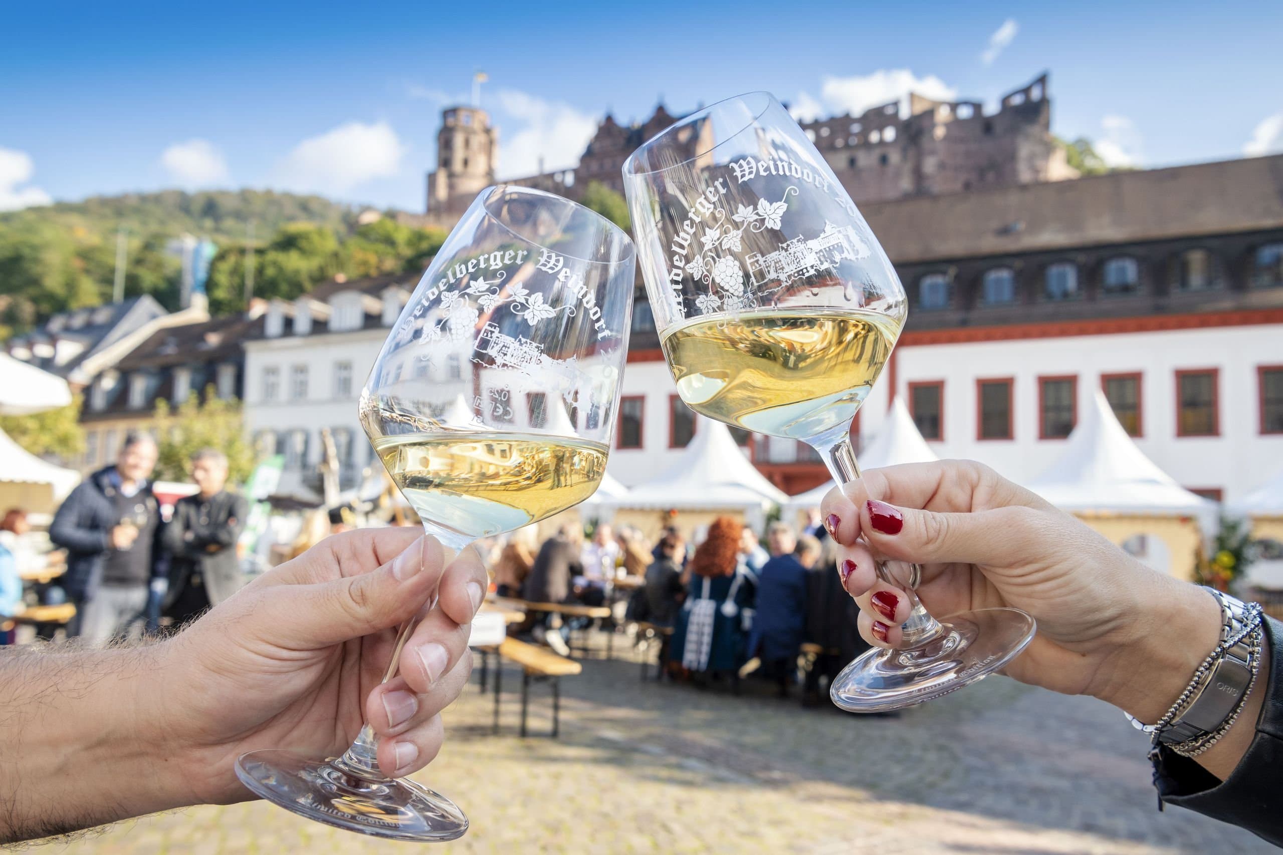 Two people clink glasses at the Heidelberg Wine Village, with Karlsplatz and Heidelberg Castle in the background.