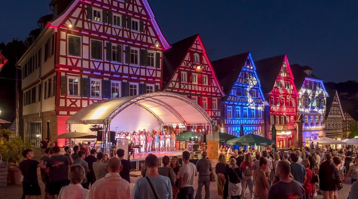 auf einem Marktplatz mit illuminierter Fachwerkkulisse stehen Menschen vor einer Bühne und tanzen