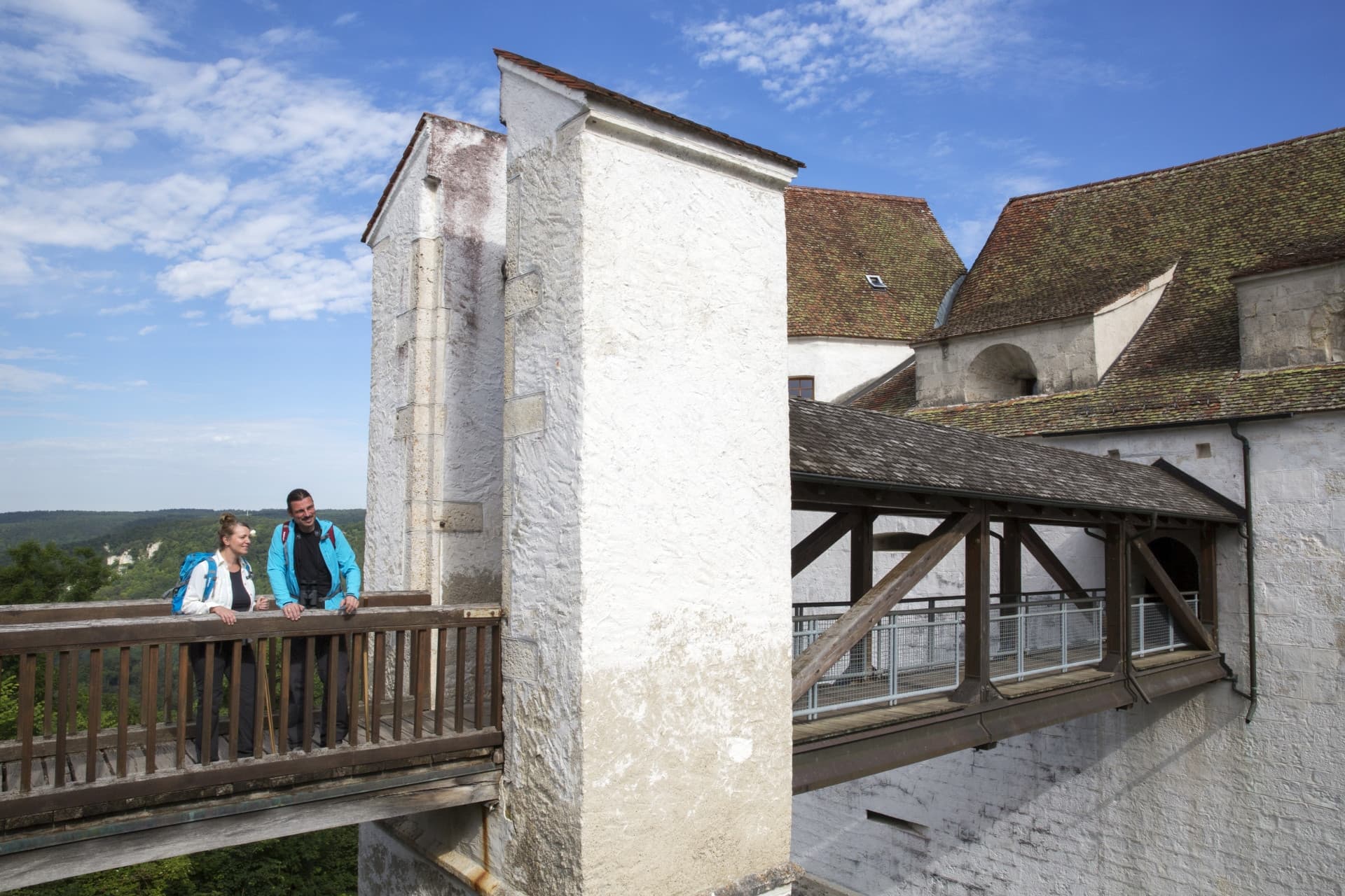 Ein Mann und eine Frau stehen auf einer Holzbrücke die zum Eingang einer mittelalterlichen weiß getünchten Burg führt
