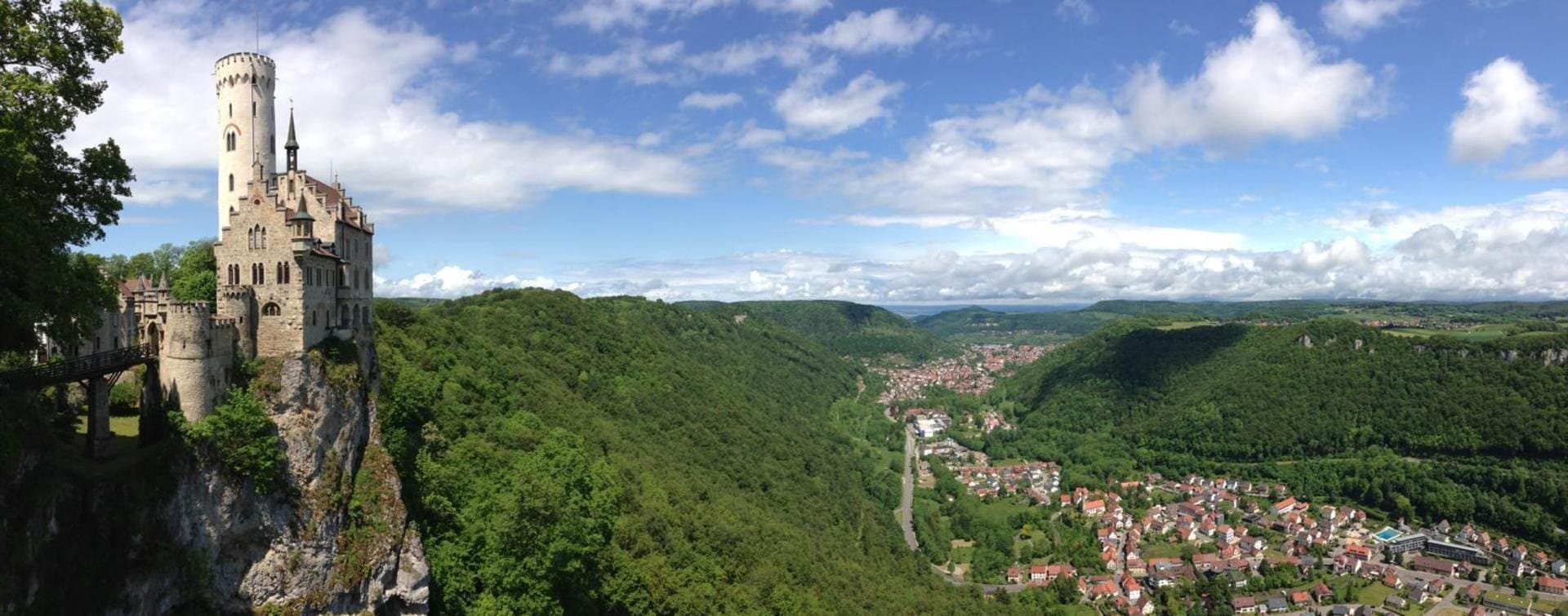 A castle with a large white tower, whose facade is ornately decorated, stands on a rock. In front of it is a bridge that leads to the other side. On the right half of the picture is a view over a valley with a city. Around it is forest.