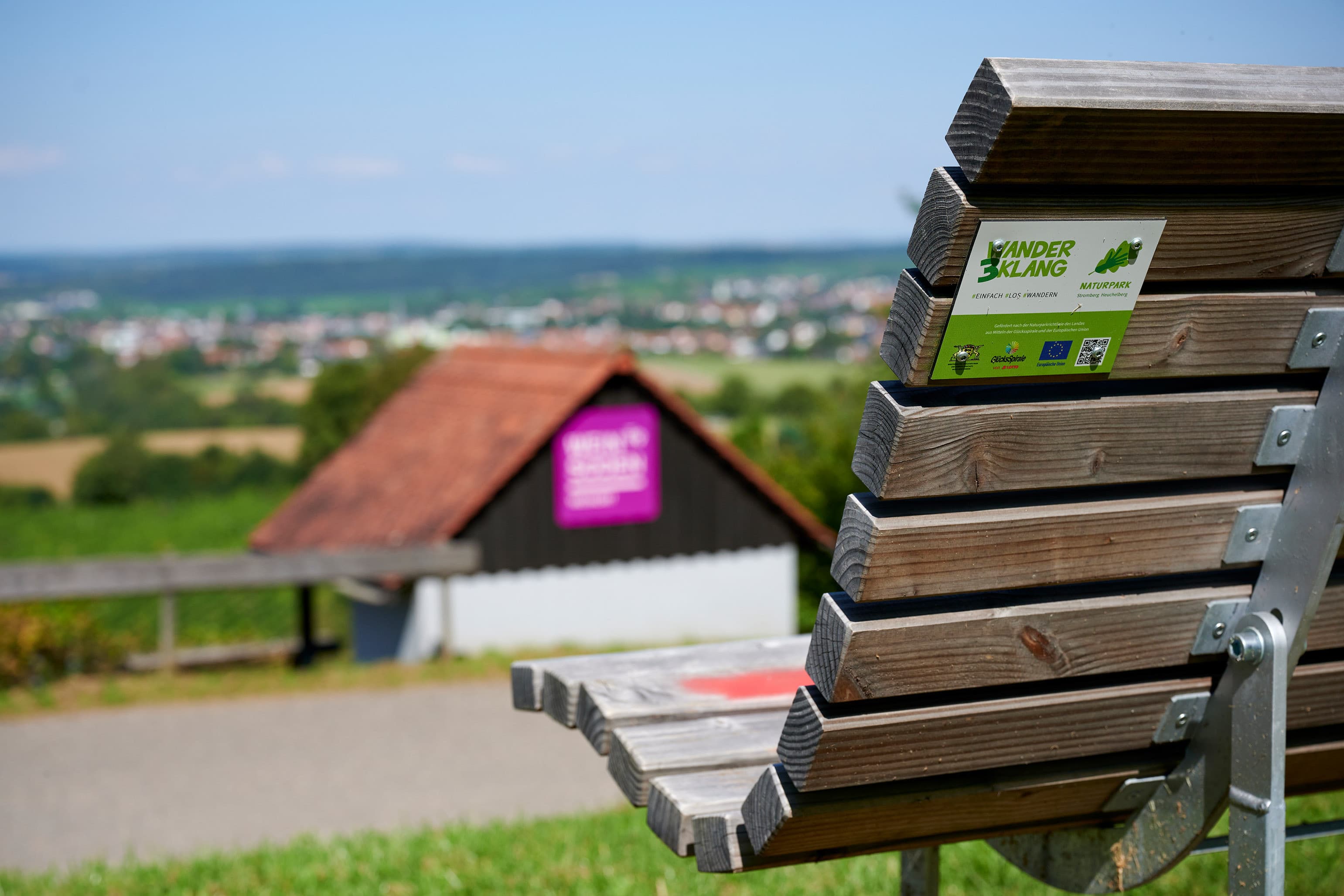 Holzbank mit Rückenlehne im Vordergrund, darauf ein Aufkleber "KinderErlebnisWeg", im Hintergrund ein weiter Ausblick über die Hügellandschaft und ein kleines Häuschen mit lila Infotafel – aufgenommen bei sommerlichem Wetter in Oberderdingen.