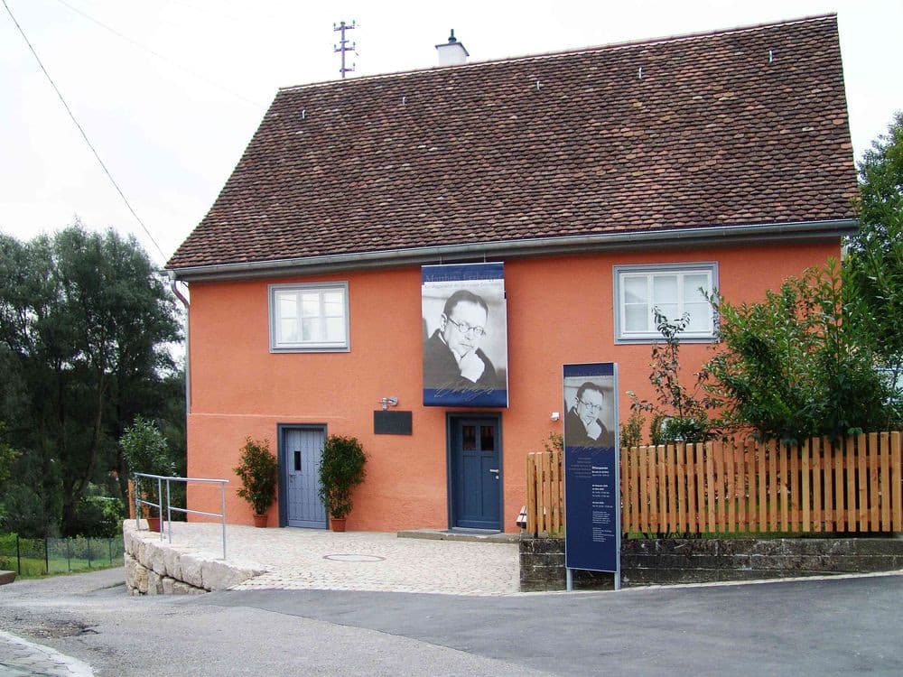 An einem orangenes Wohnhaus mit kleinem Garten davor und blauen Türen hängt ein Banner mmit einer Person darauf. Auf einem Schild an der Hofeinfahrt ist die Person ebenfalls abgebildet und es stehen weitere Infos darauf.