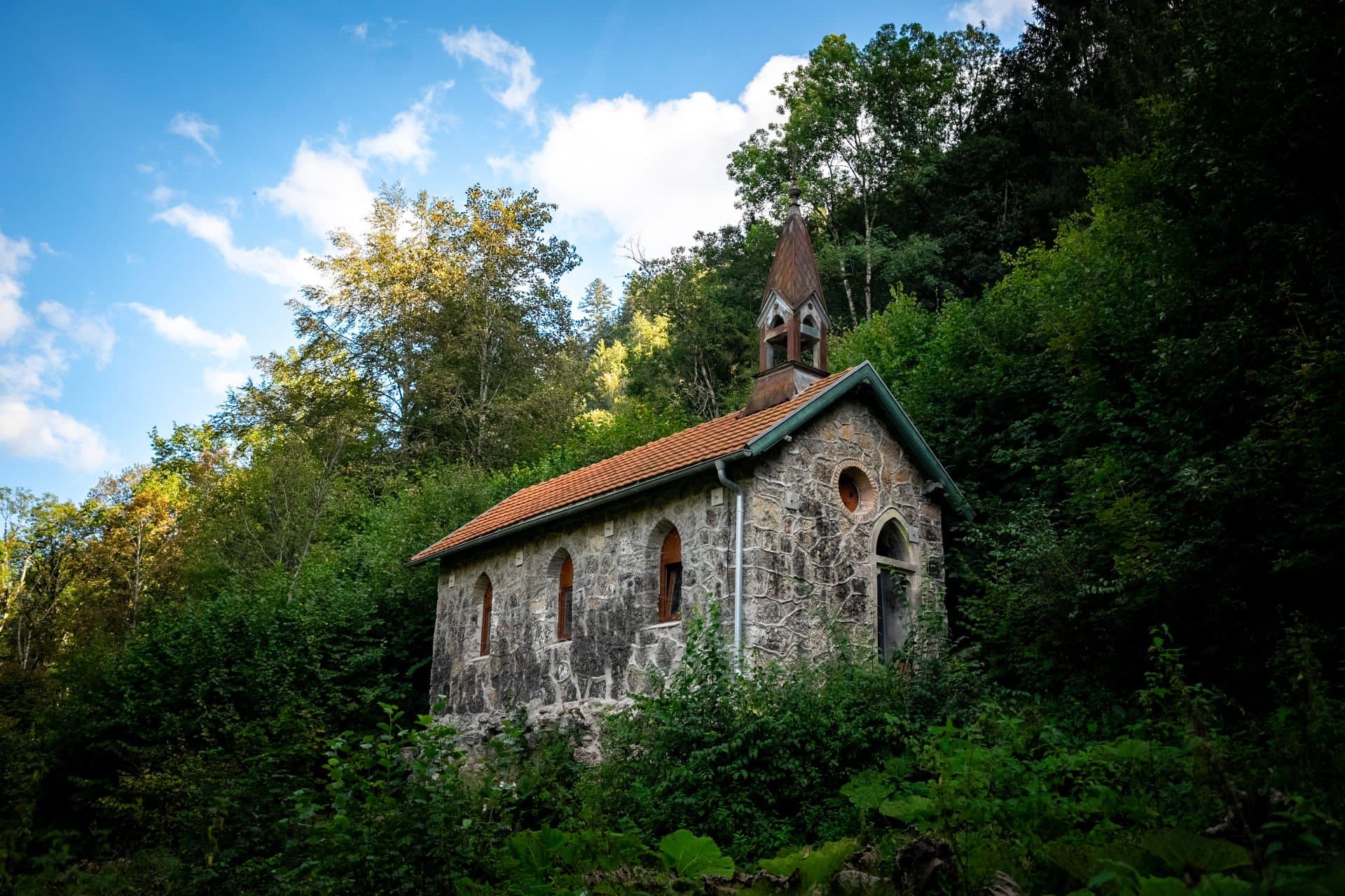 Eine steinerne Kapelle mit orangenem Dach steht an einem Berg. Rund um die Kapelle herum sind Sträucher und Bäume.