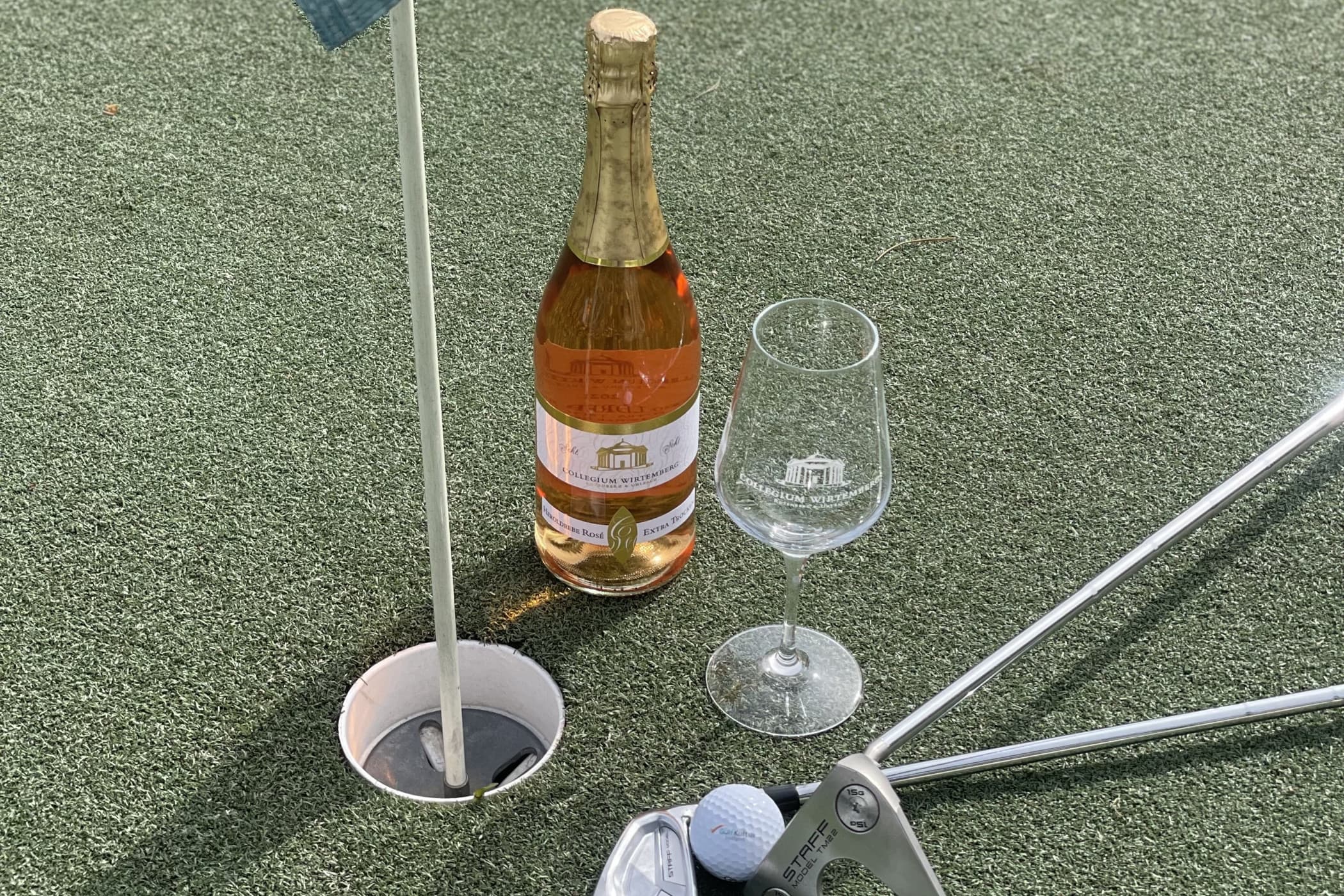 Eine Sektflasche steht auf Golfrasen neben einem Loch und einem Golfschläger