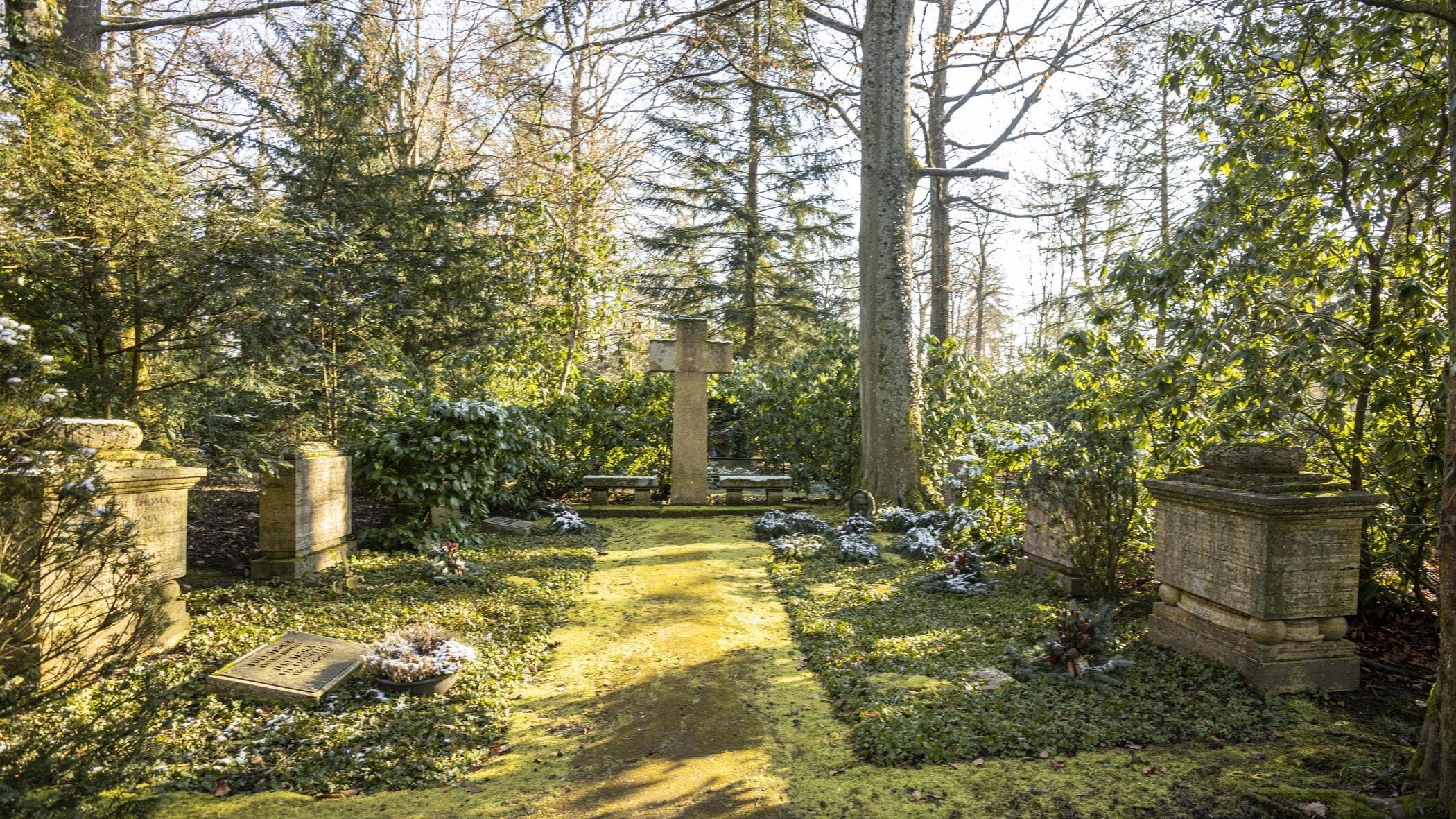 Blick ins Grün. Ein Grasstreifen führt auf ein großes Steinkreuz zu. Rechts und links des Pfads befinden sich weitere Grabsteine. Im Hintergrund stehen große Laub- und Nadelbäume.