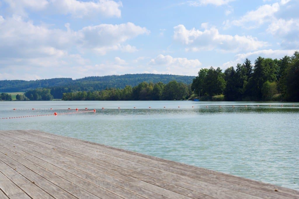 Blick vom Holzsteg auf den blauen Illmensee, hinter dem sich Bäume und der Himmel erheben.