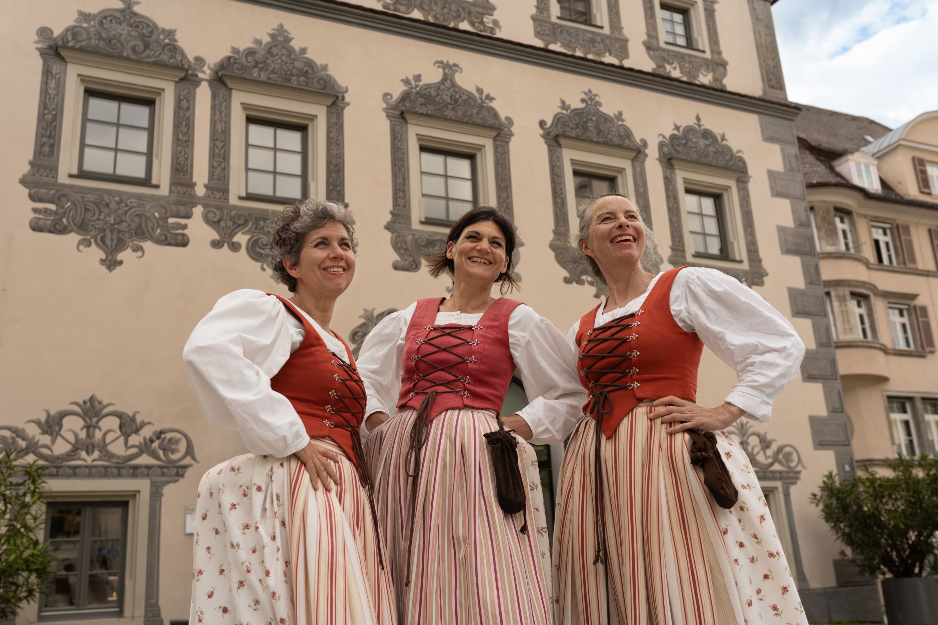 Three women in traditional costumes in front of a historic building.  The three actresses meet in front of the Lederhaus.