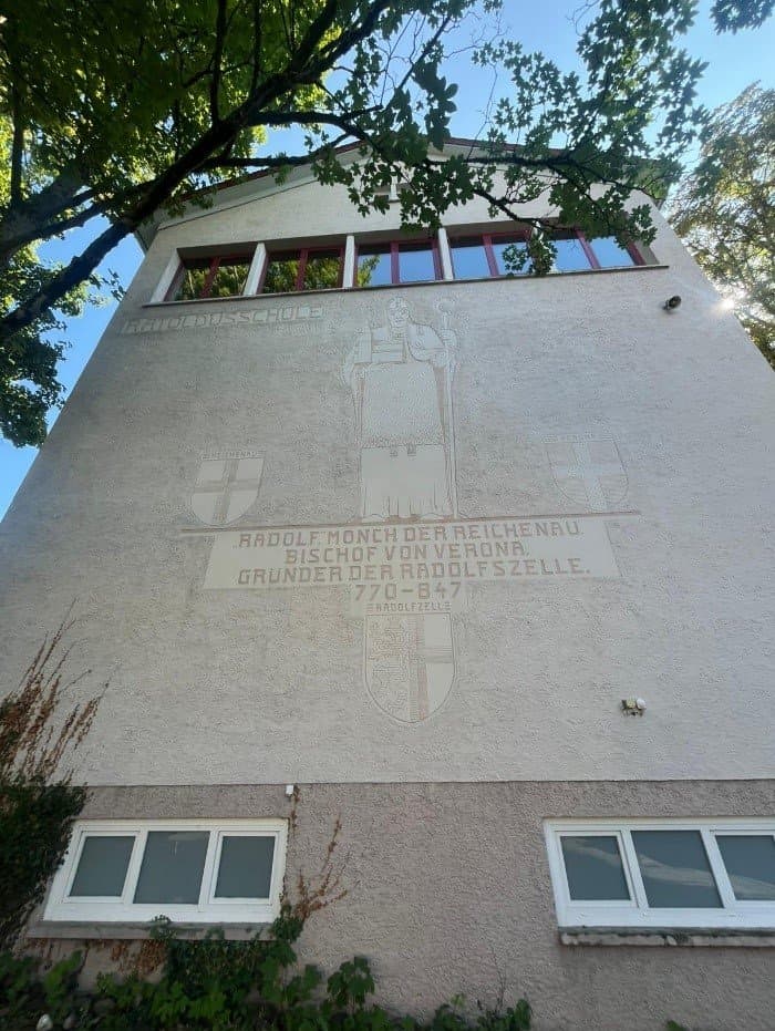 Das Bild zeigt die Ratoldus-Schule in Radolfzell.  Fassade mit Fenstern und Ratoldus; links das Wappen Reichenau, rechts Verona. Schriftzug: „Radolf, Mönch der Reichenau, Bischof von Verona. Gründer der Radolszelle. 770–847.“ Unten Radolfzeller Wappen.