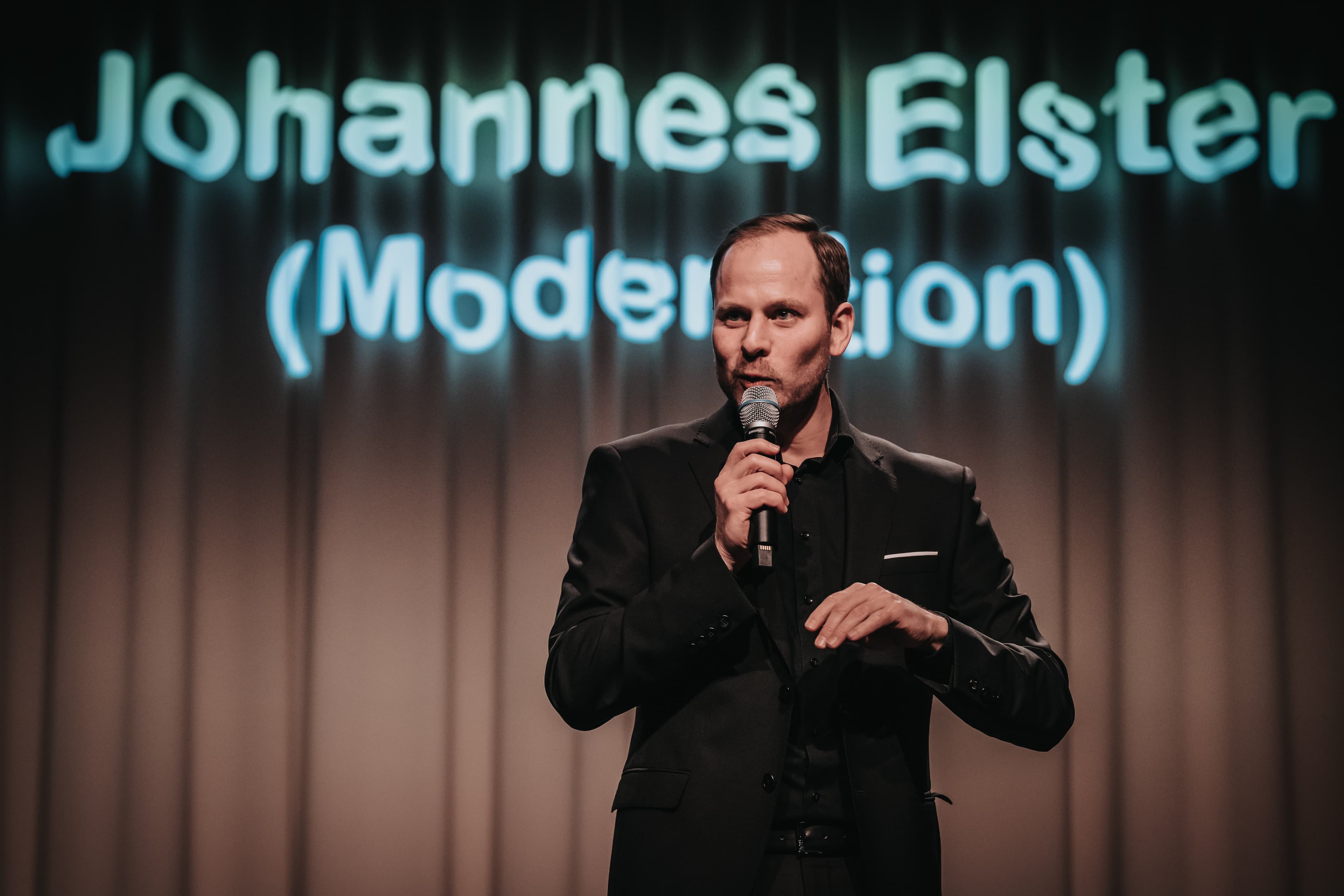Auf dem Bild ist ein Mann mit Mikrofon zu sehen.  Er steht vor einer Leinwand mit dem Schriftzug "Johannes Elster (Moderation)".