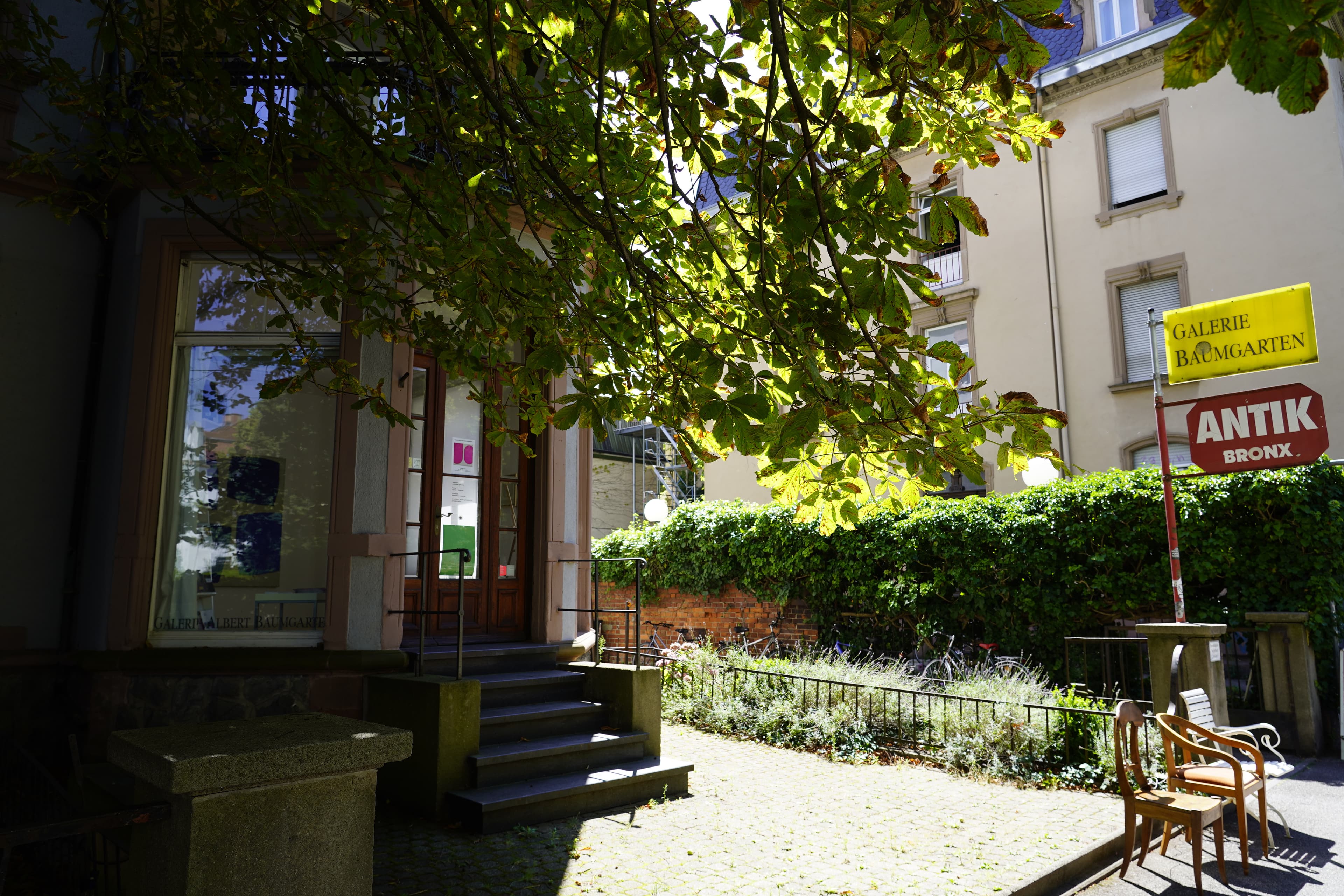Eingang zur Galerie Baumgarten mit Schildern und Stühlen im Freien.