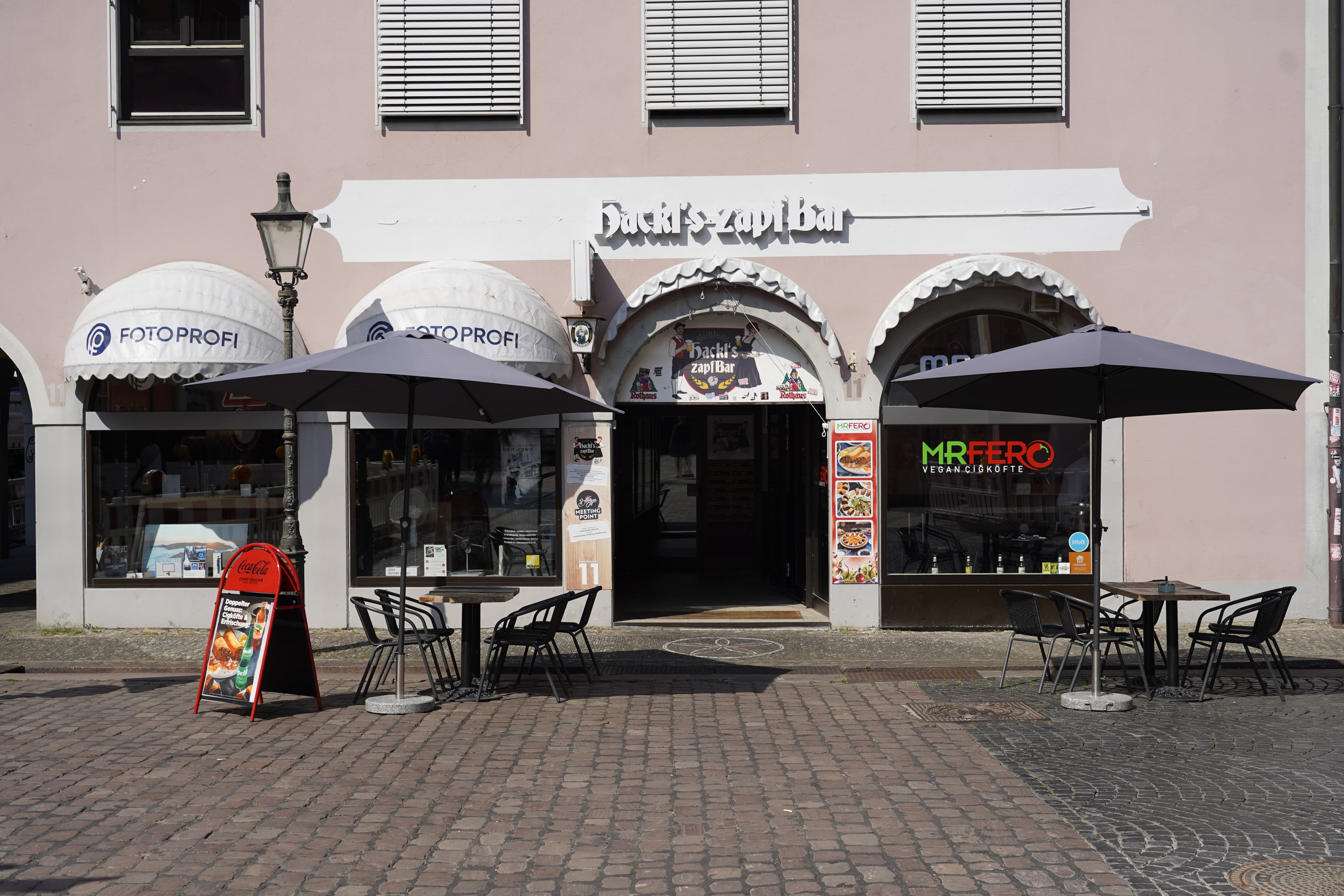 Außenansicht von Hackl's Zapfbar mit Tischen und Sonnenschirmen auf dem Gehweg.