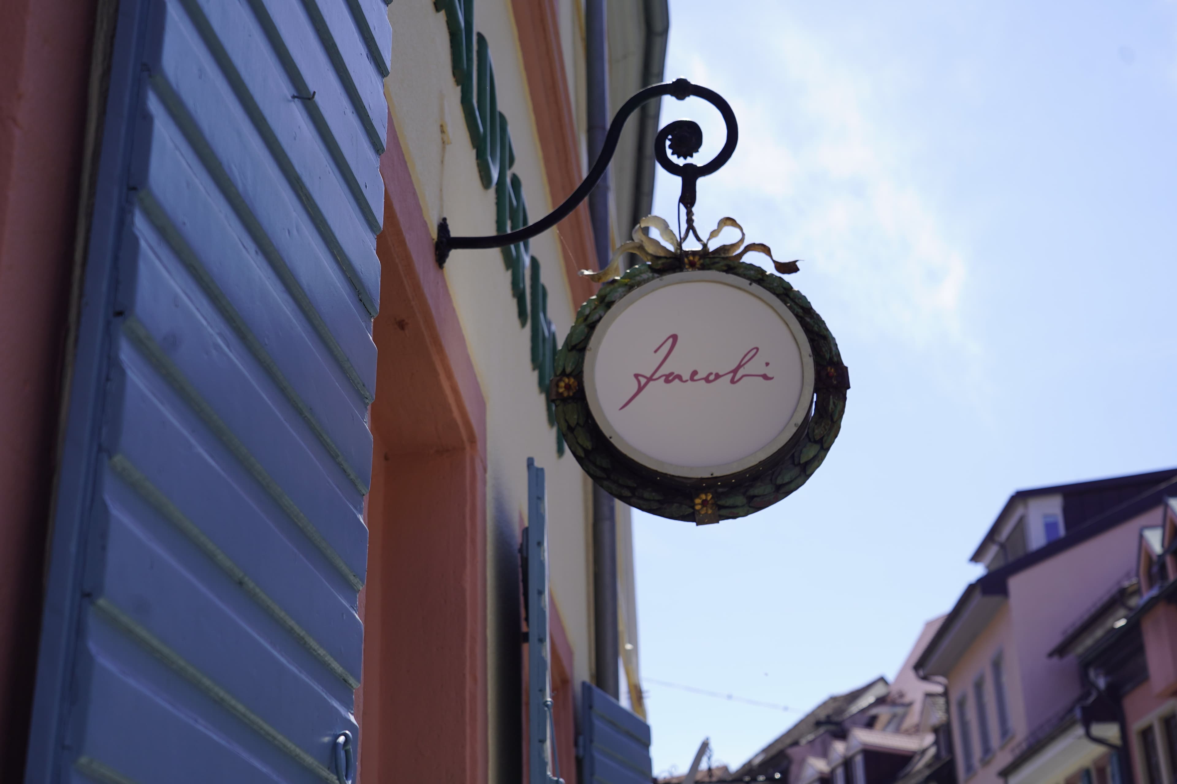 Ein Schild mit der Aufschrift 'Jacobi' hängt an einer Hauswand mit blauen Fensterläden.