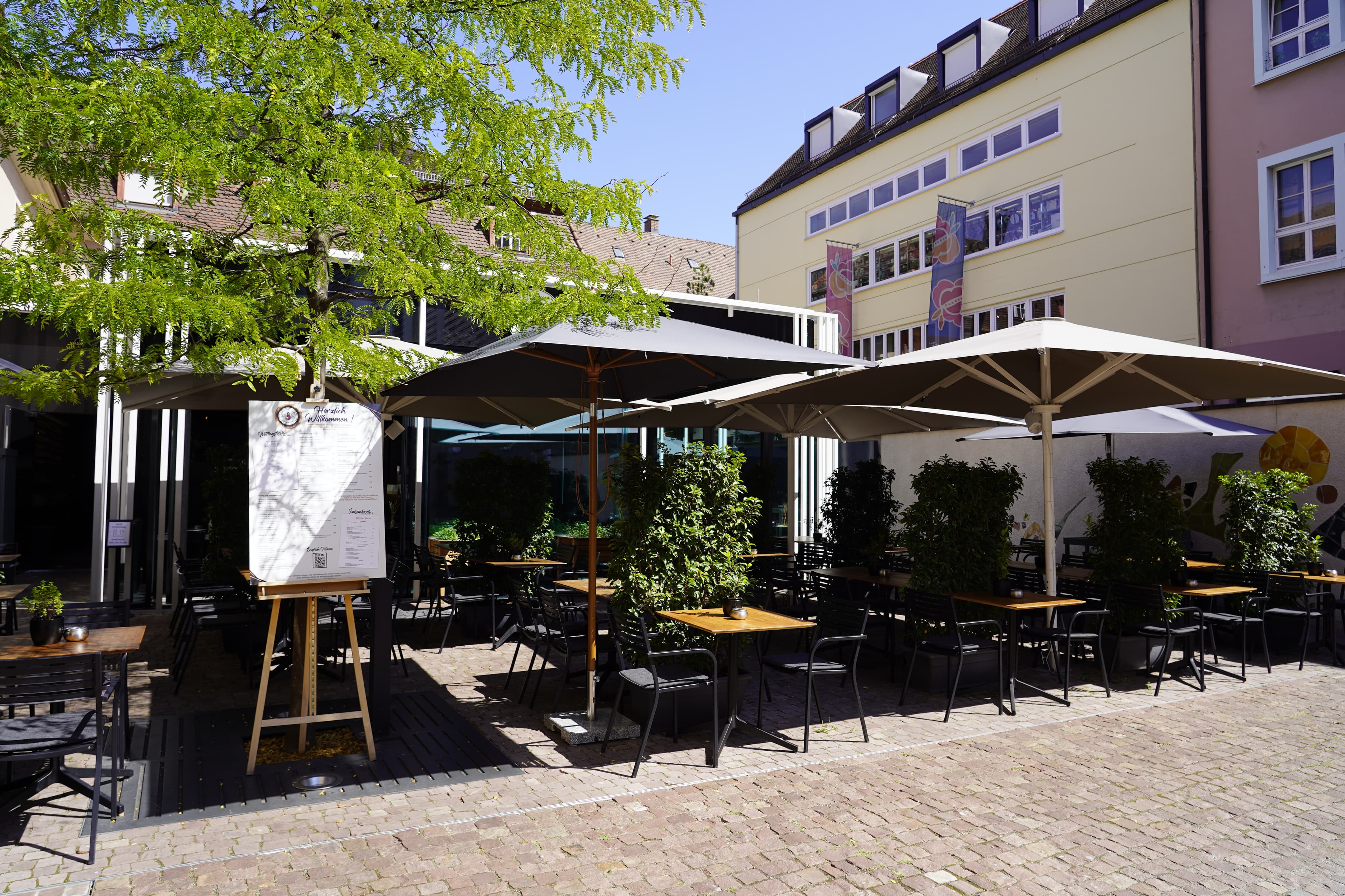 Außenbereich eines Cafés mit Tischen, Stühlen und Sonnenschirmen auf einem gepflasterten Platz.
