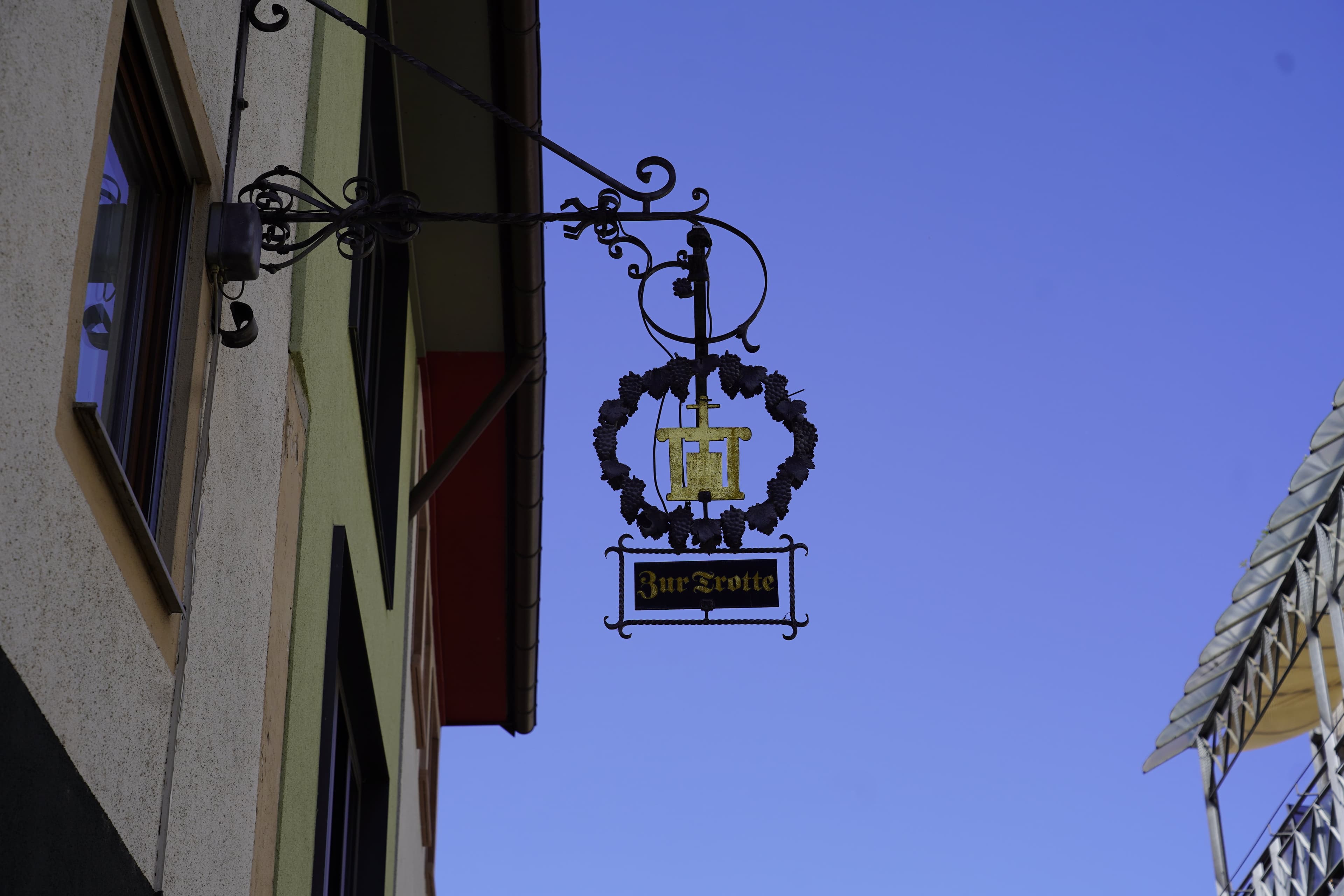 Ein dekoratives Schild mit der Aufschrift 'Zur Trotte' hängt an einem Gebäude vor blauem Himmel.