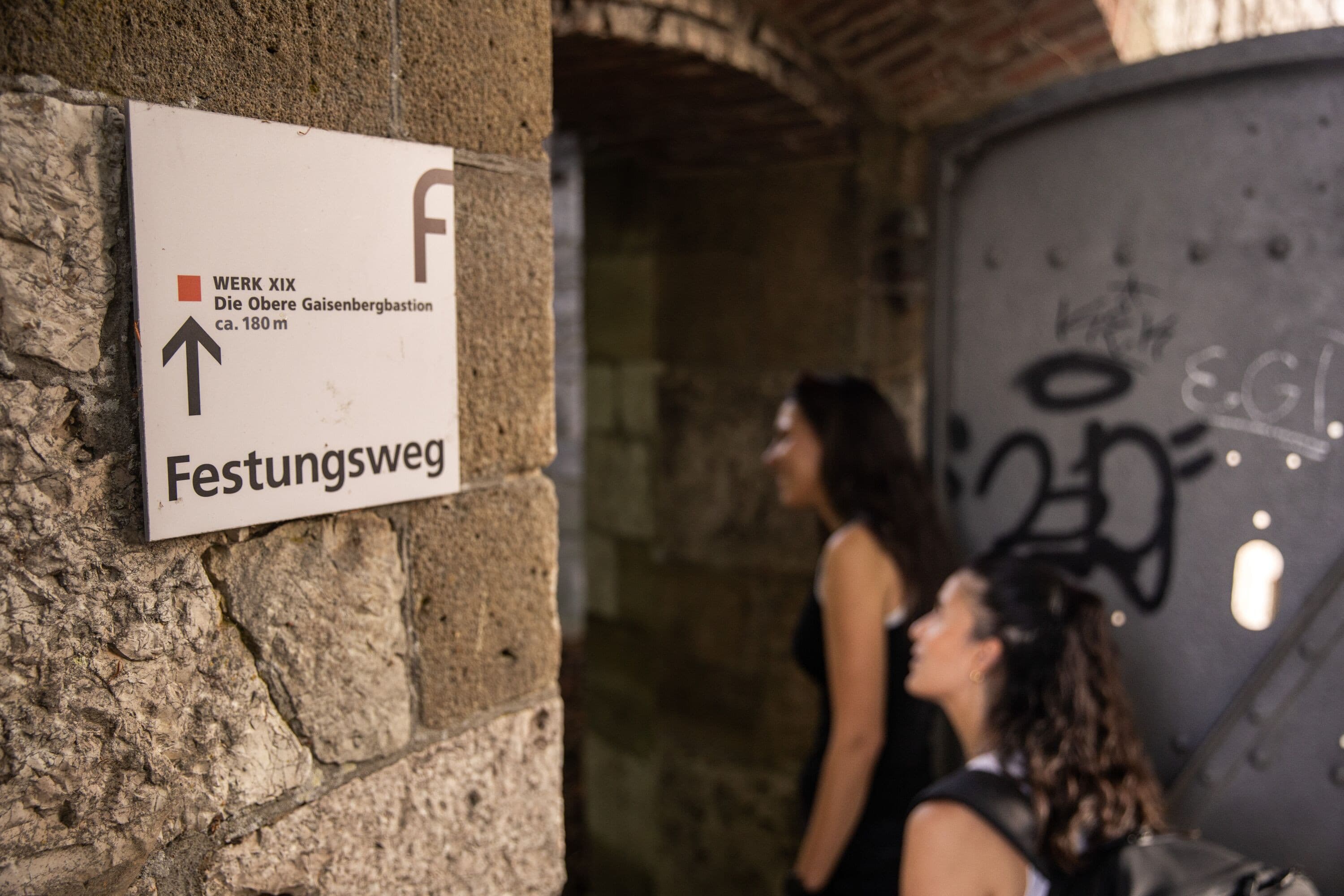 Informationsschild „Festungsweg“ an einer Steinmauer, zwei Frauen gehen durch einen Torbogen.