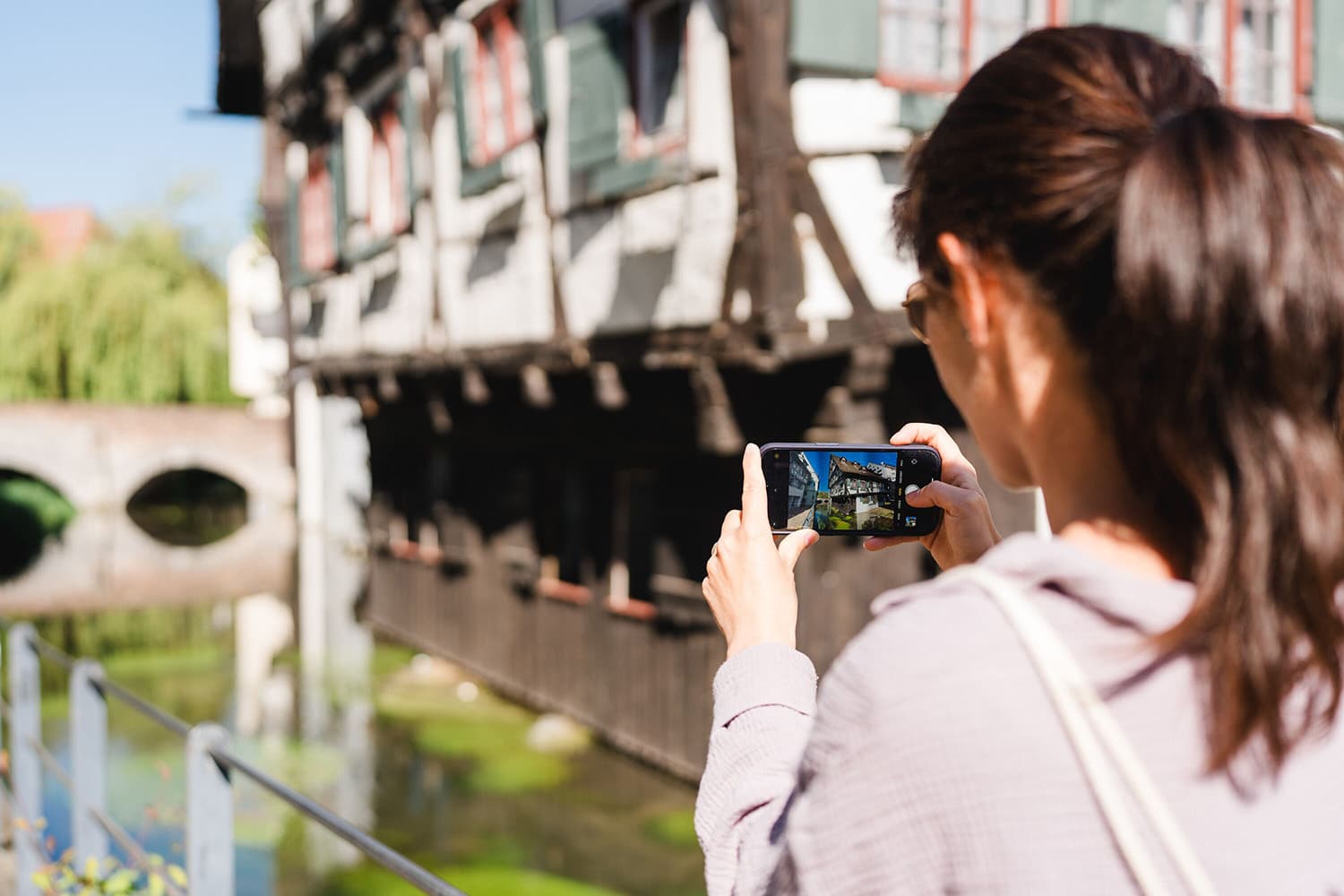 Frau fotografiert mit dem Smartphone ein schiefes Fachwerkhaus am Wasser.
