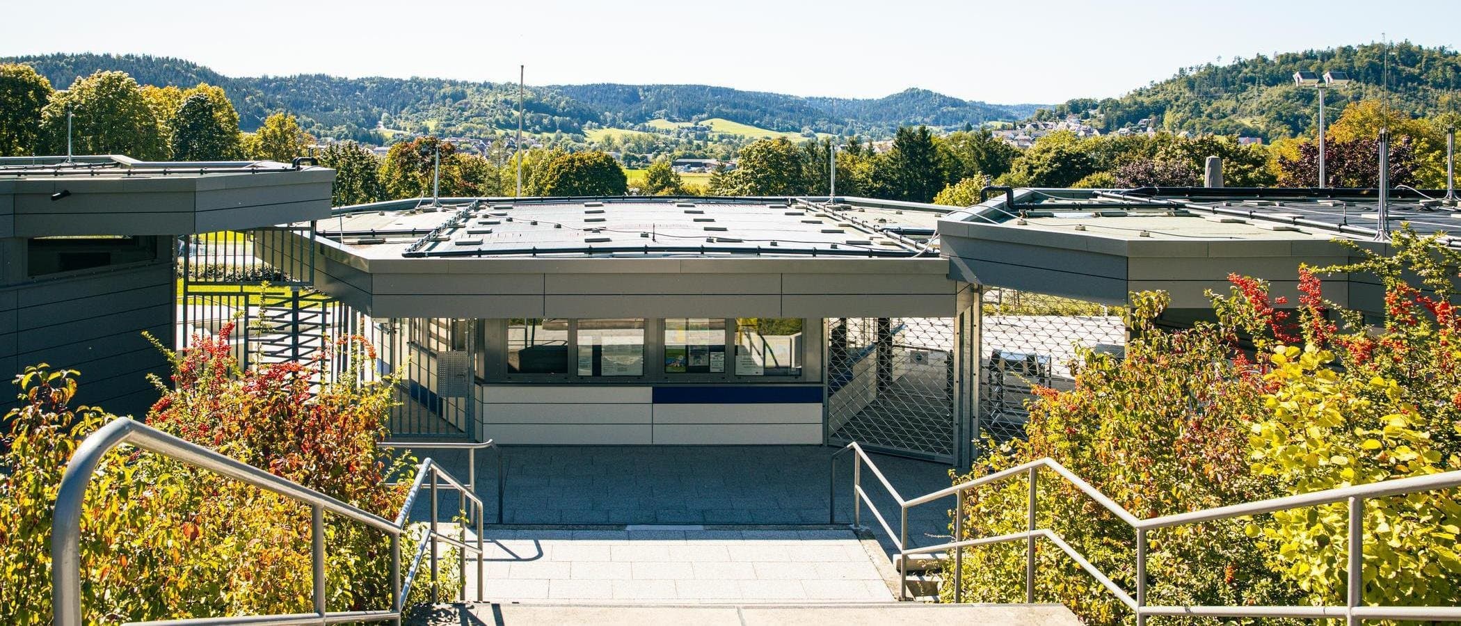 Eingangsbereich eines modernen Freibads mit grüner Umgebung und Hügeln im Hintergrund.