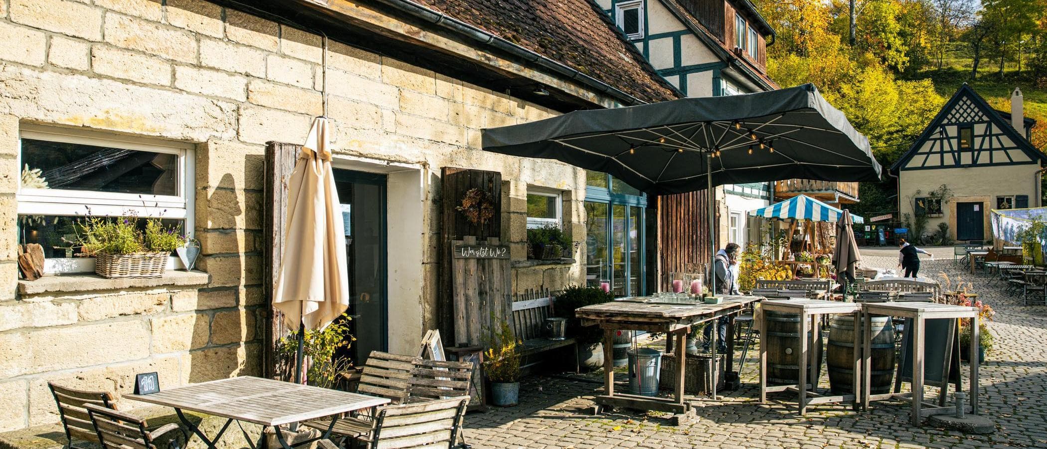 Ein gemütlicher Außenbereich eines Cafés mit Holztischen und Stühlen, umgeben von alten Gebäuden und herbstlicher Vegetation.