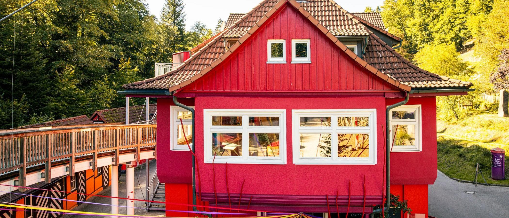 Ein rotes Gebäude mit weißen Fenstern und einem Holzsteg im Vordergrund, umgeben von Bäumen.