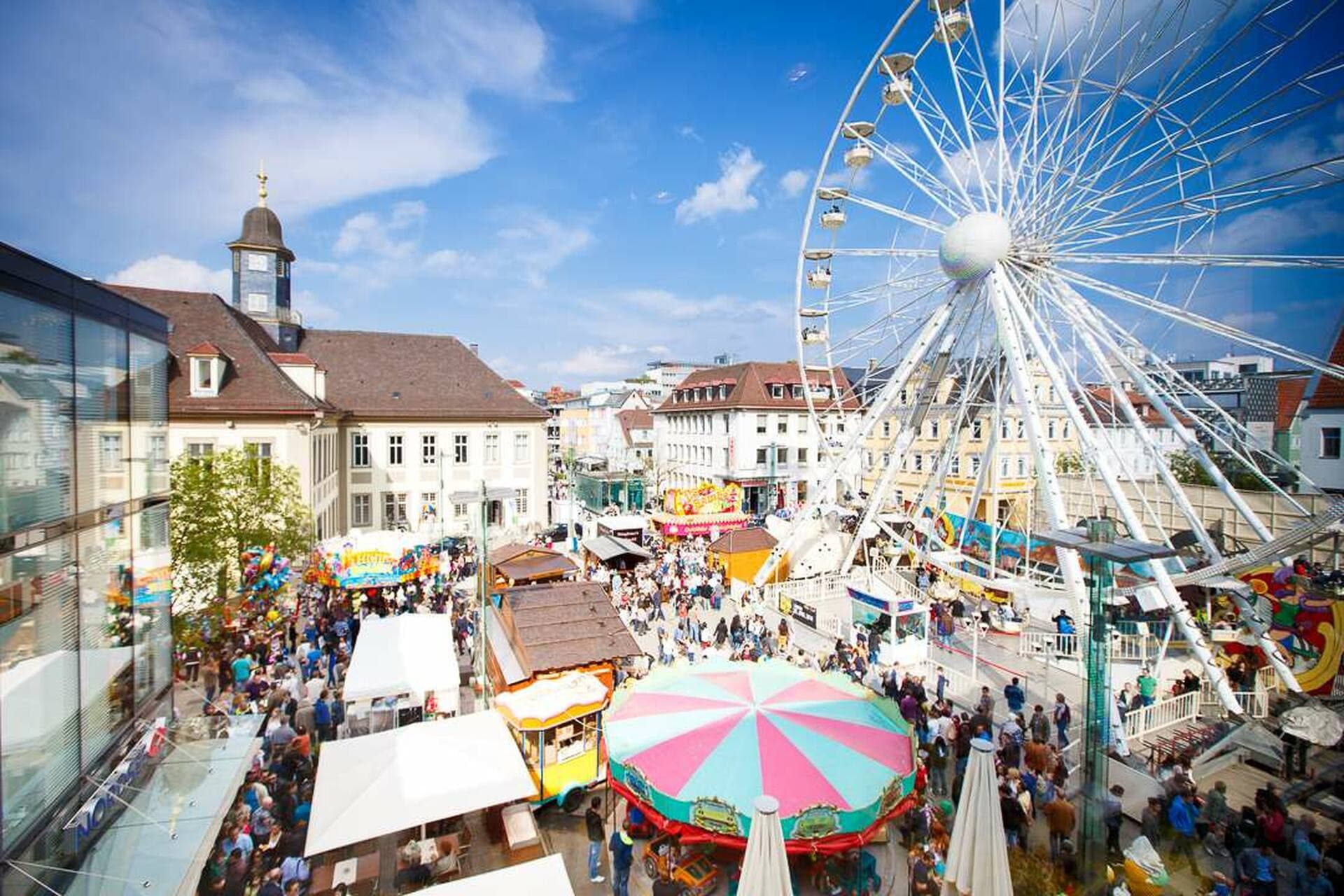 Ein belebter Platz in Göppingen mit einem großen Riesenrad und vielen Menschen bei sonnigem Wetter.