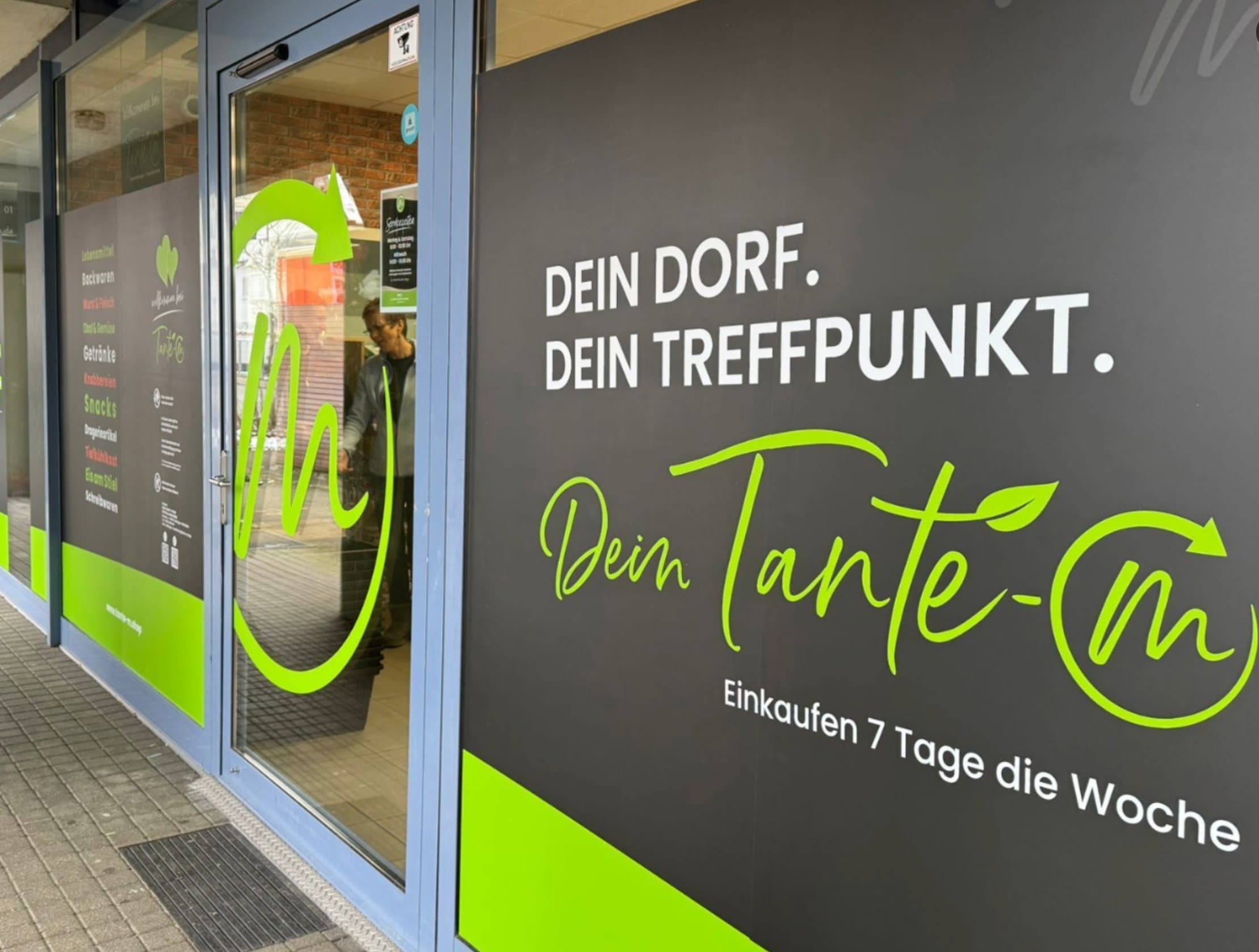 Tante-M Laden mit grünem Logo und Werbeslogan 'Dein Dorf. Dein Treffpunkt.' Einkaufen 7 Tage die Woche.