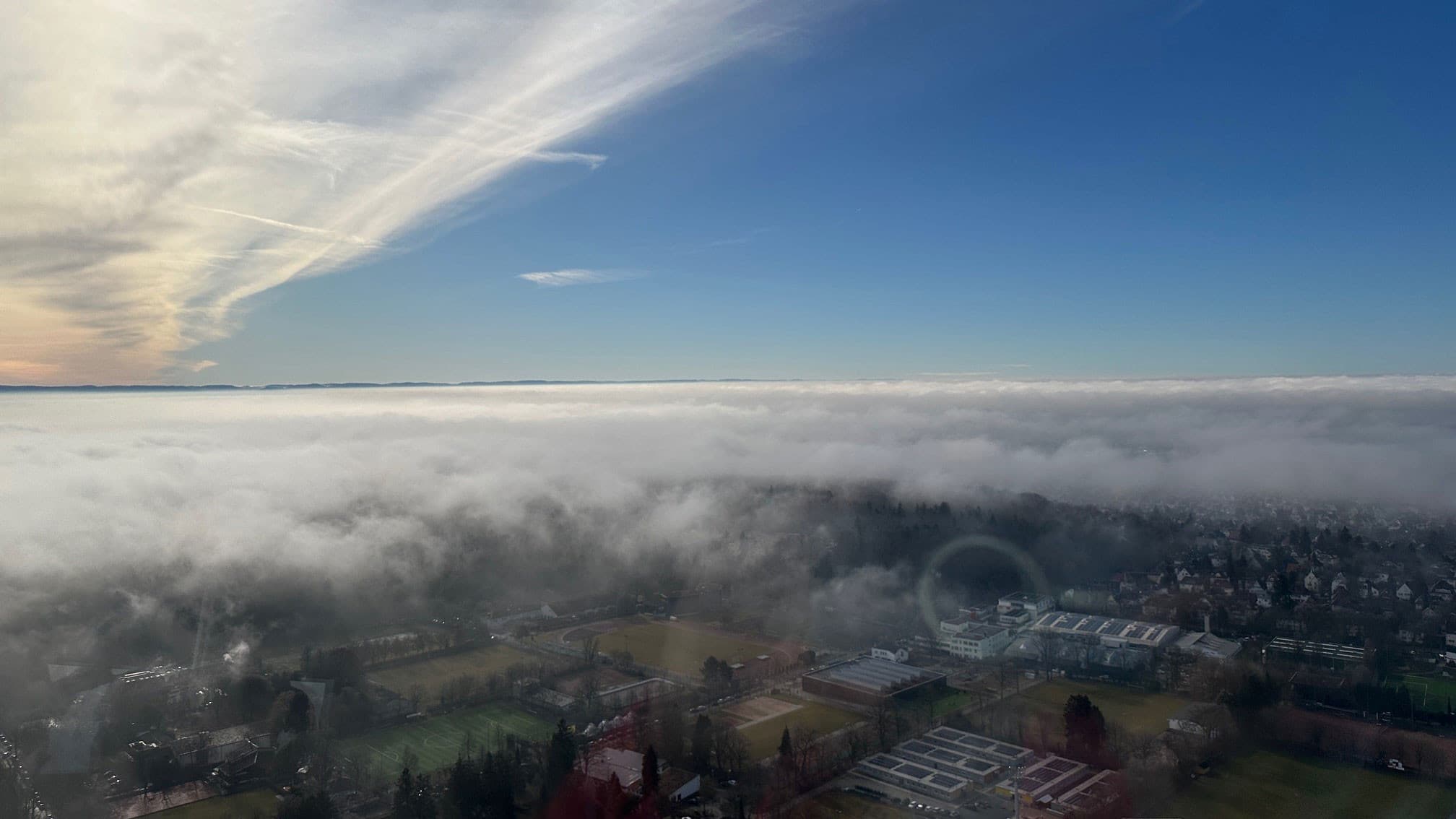 Blick vom Fernsehturm auf eine Stadt unter einer Wolkendecke mit blauem Himmel darüber.