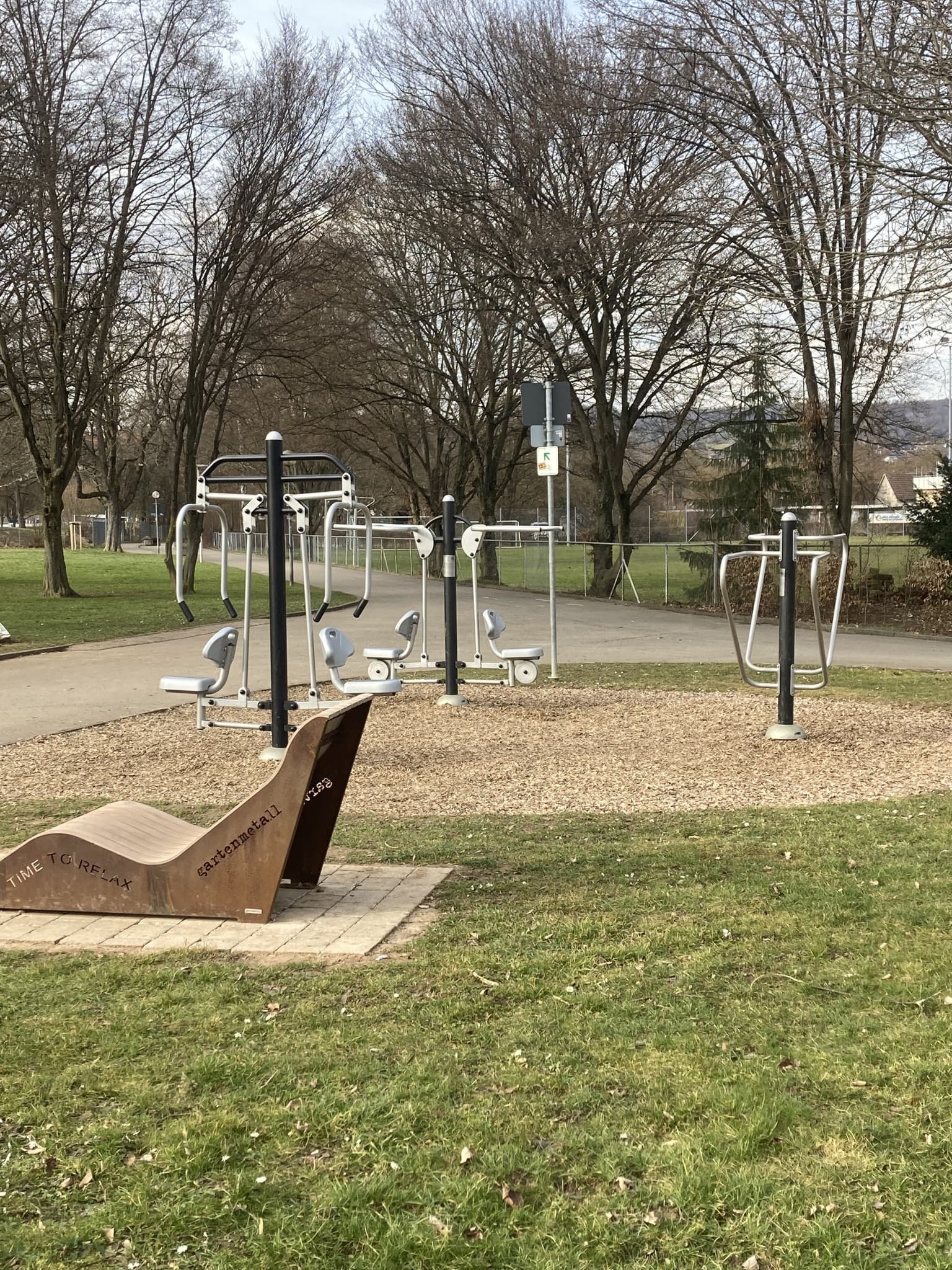 Ein Outdoor-Fitnessbereich mit verschiedenen Trainingsgeräten und einer Holzbank in einem Park.