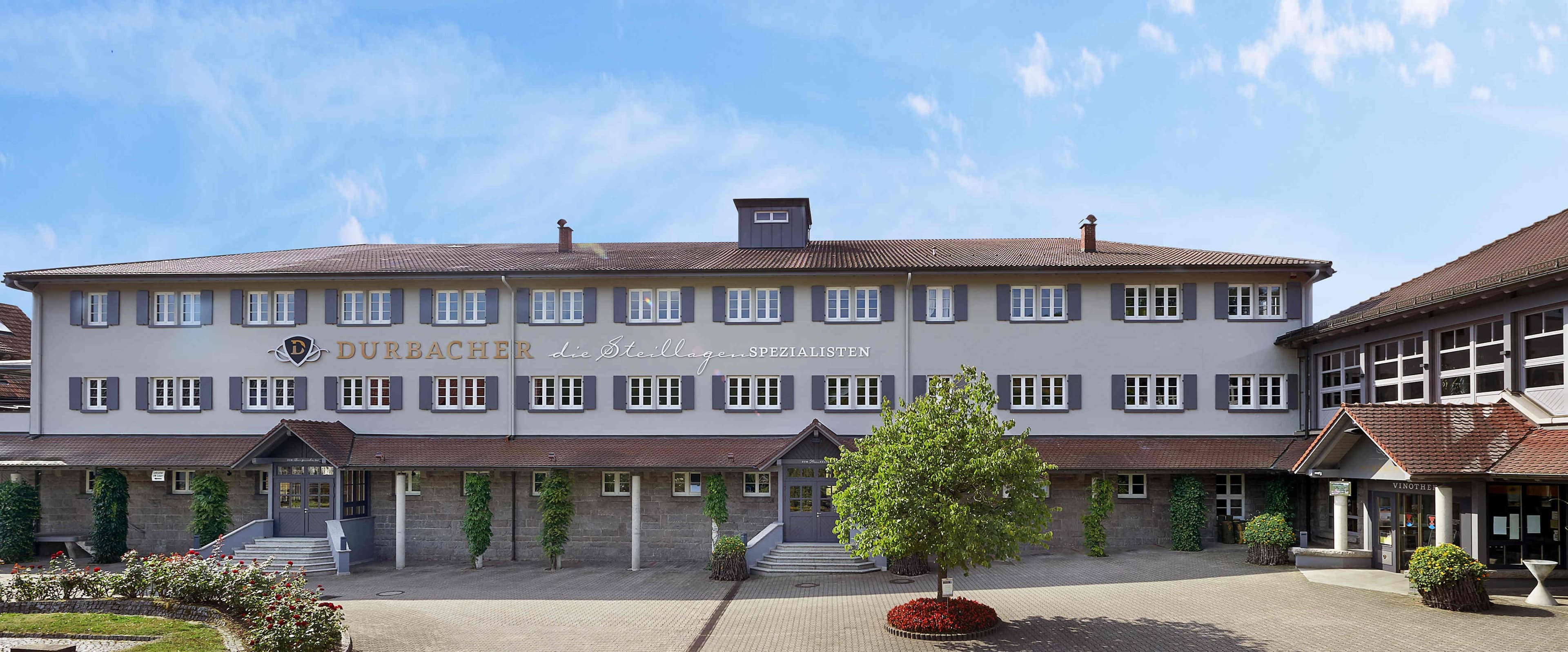Langgestrecktes Gebäude mit vielen Fenstern und goldenem Schriftzug und schöner Bepflanzung im Vordergrund