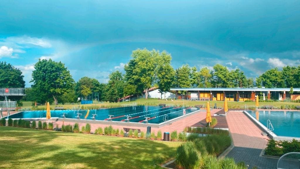Es sind zwei Schwimmbecken zu sehen mit abgetrennten Bahnen und den Sprungtürmen und der Liegewiese vor und hinter den Becken.