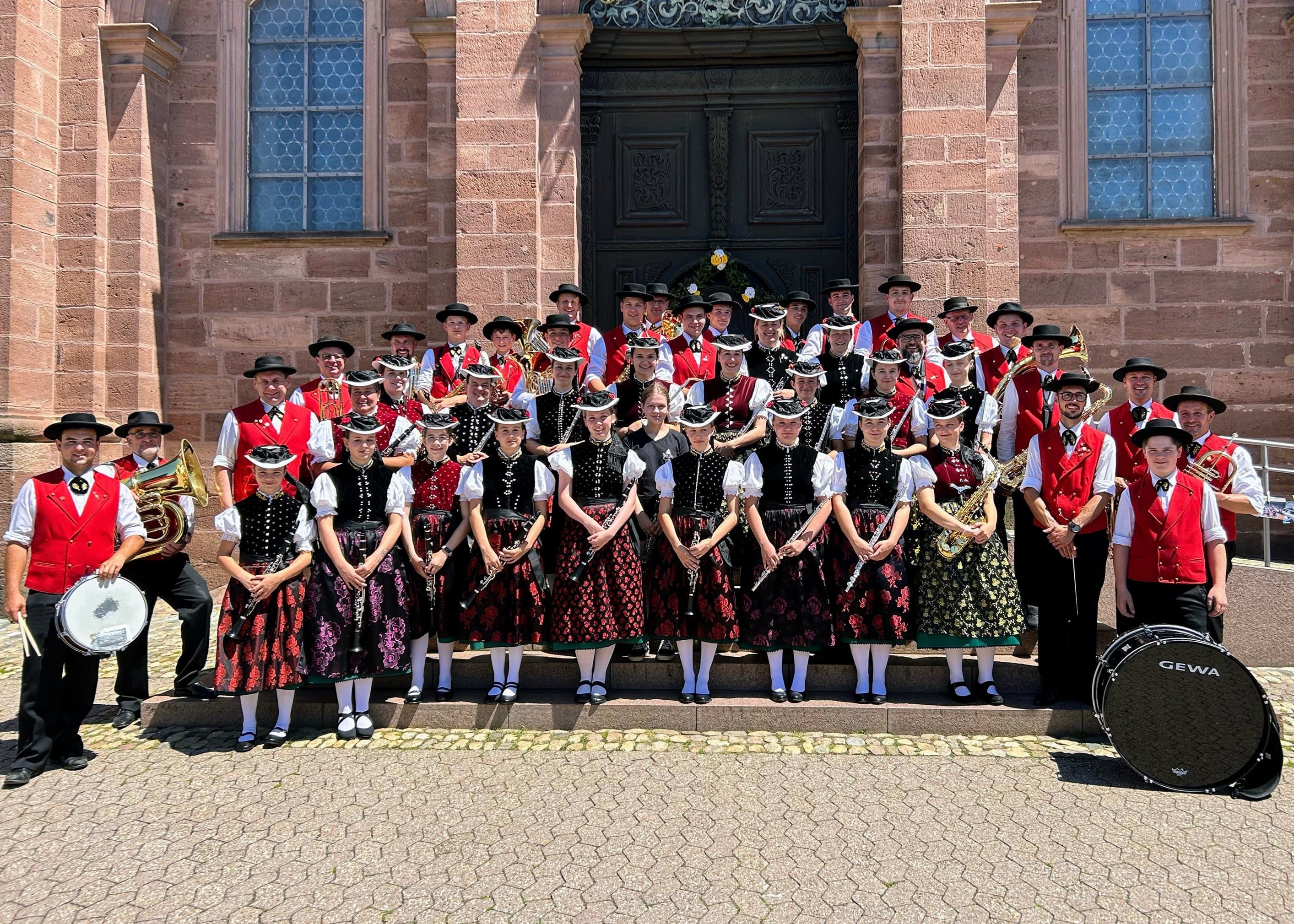 Musiker:innen in festlicher Schwarzwälder Tracht mit ihren Instrumenten vor dem Kirchenportal.