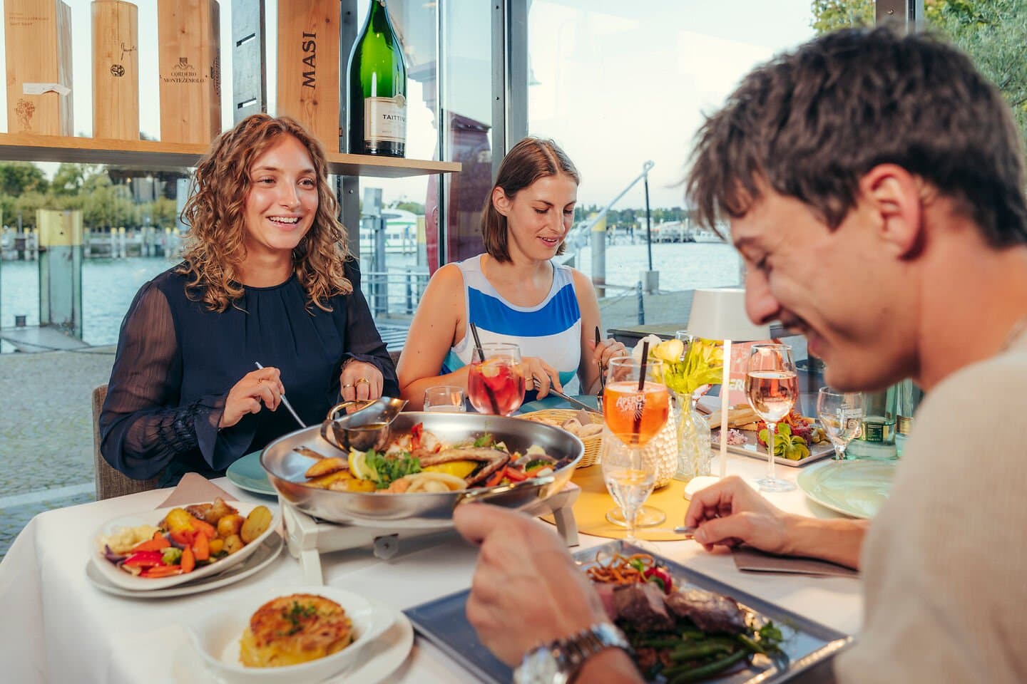 Zwei Frauen und ein Mann sitzen gemeinsam am Tisch. Sie trinken Aperitifs und Essen Fisch frisch aus er Pfanne, ein Fleischgericht sowie Beilagen.