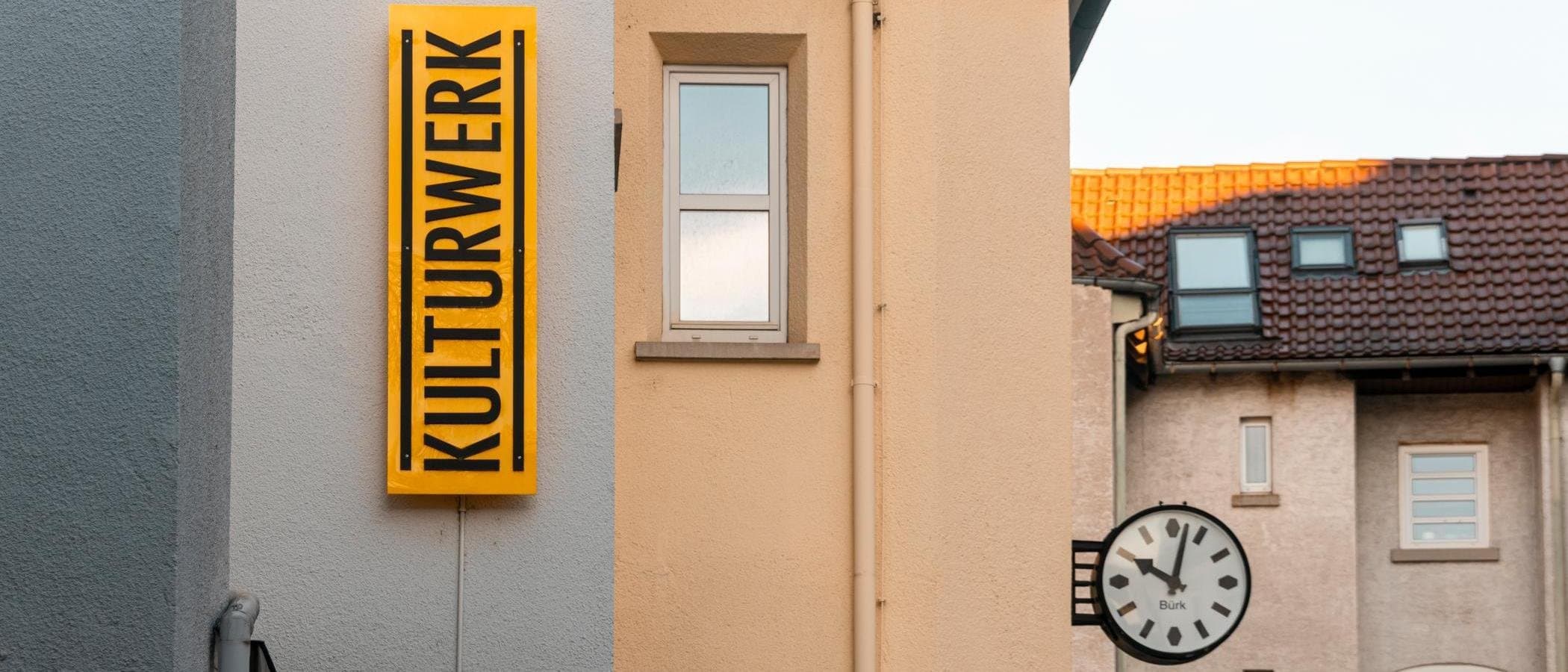 Gelbes Schild mit der Aufschrift 'KULTURWERK' an einer Hauswand, daneben ein Fenster und eine Uhr.