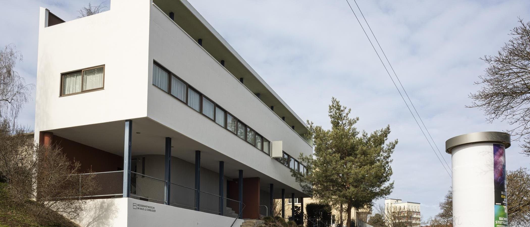 Modernes Gebäude mit weißer Fassade und großen Fenstern, umgeben von Bäumen und einem Plakatständer.
