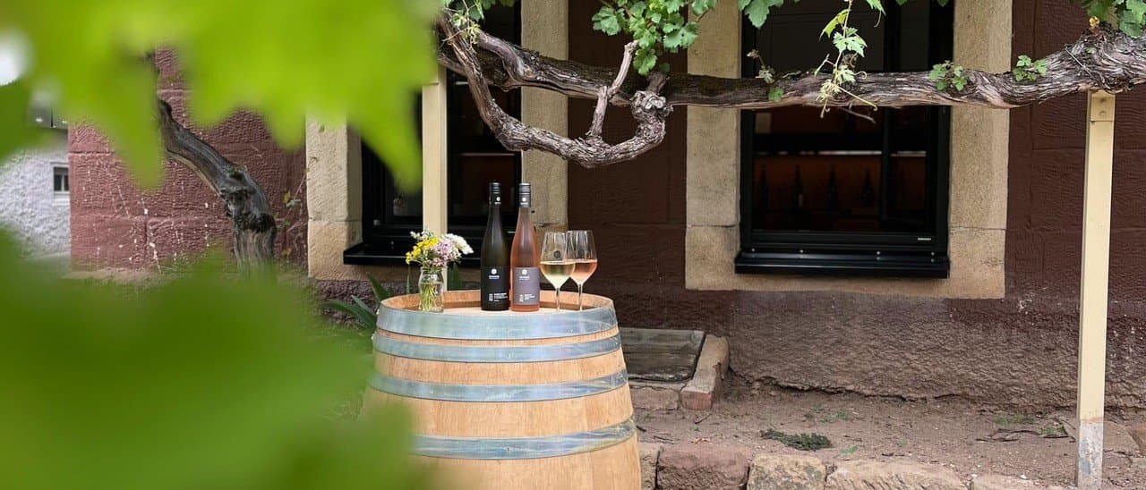 A wine barrel serves as a table, with two bottles of wine and glasses on top, surrounded by vines in front of a rustic building.