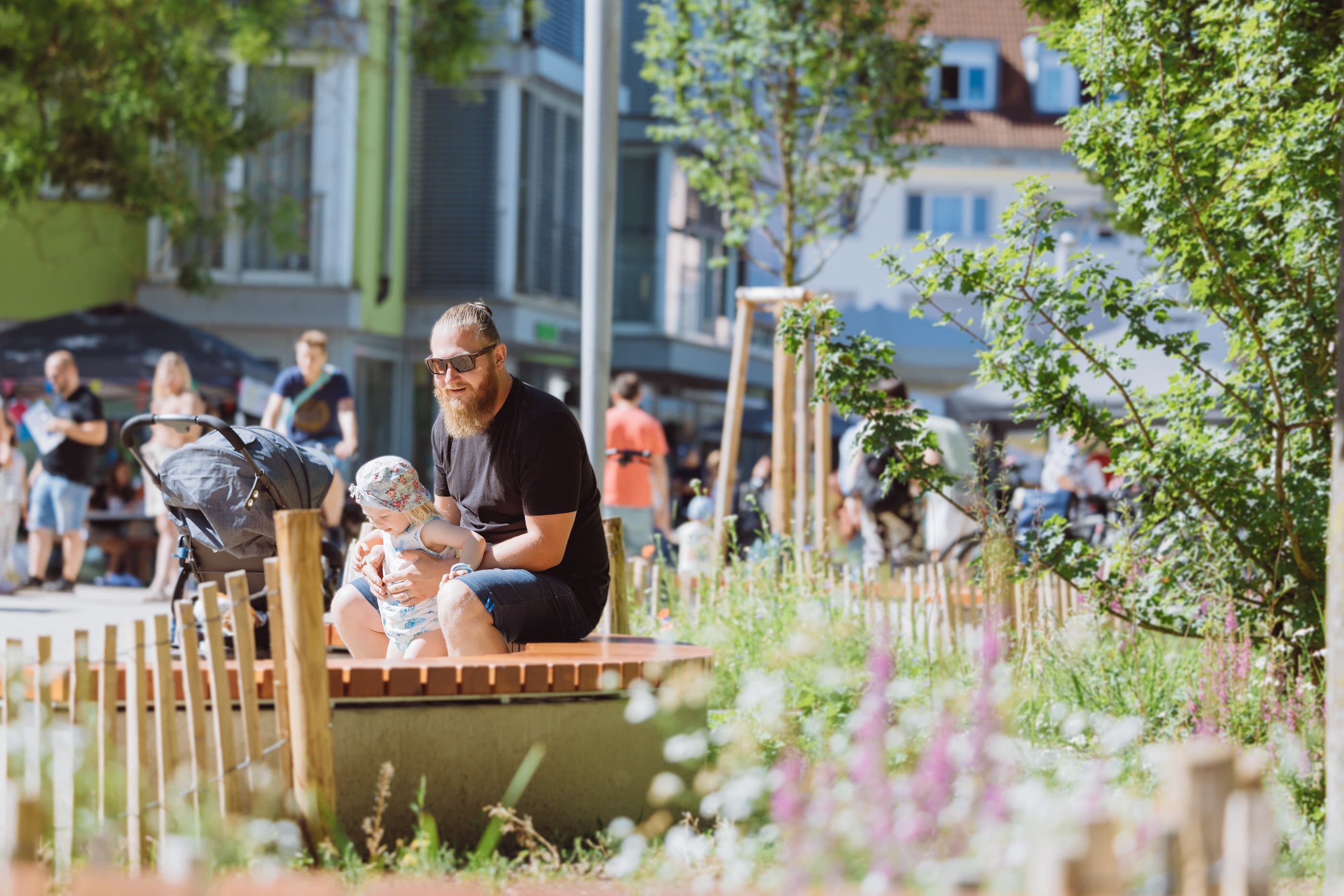 A man with a child on a wooden bench in the park, with people and buildings in the background.