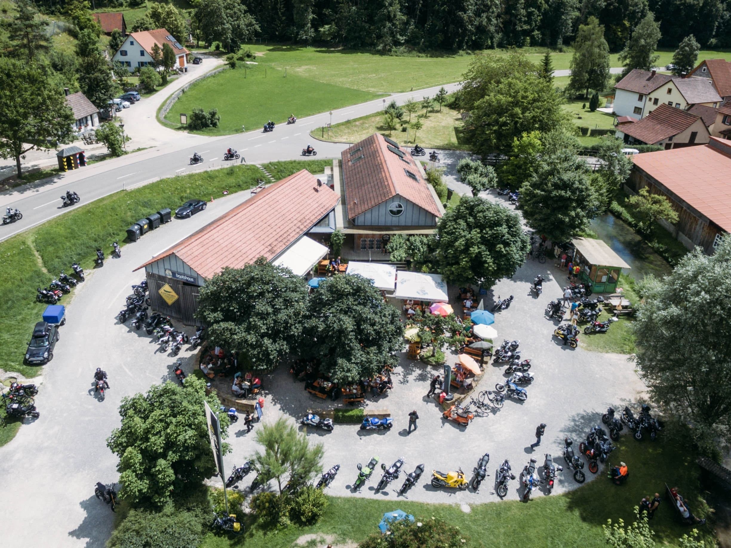 Bootshaus von oben. Auf dem Parkplatz stehen  Personen und Motorräder.
