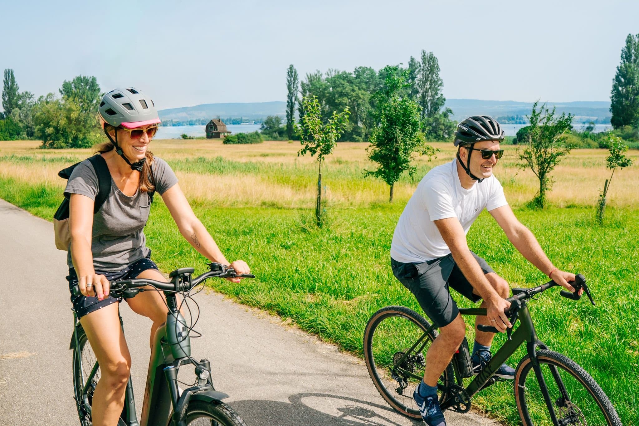 Ein Pärchen radelt auf deinem Radweg zwischen Wiesen, im Hintergrund sieht man den Bodensee