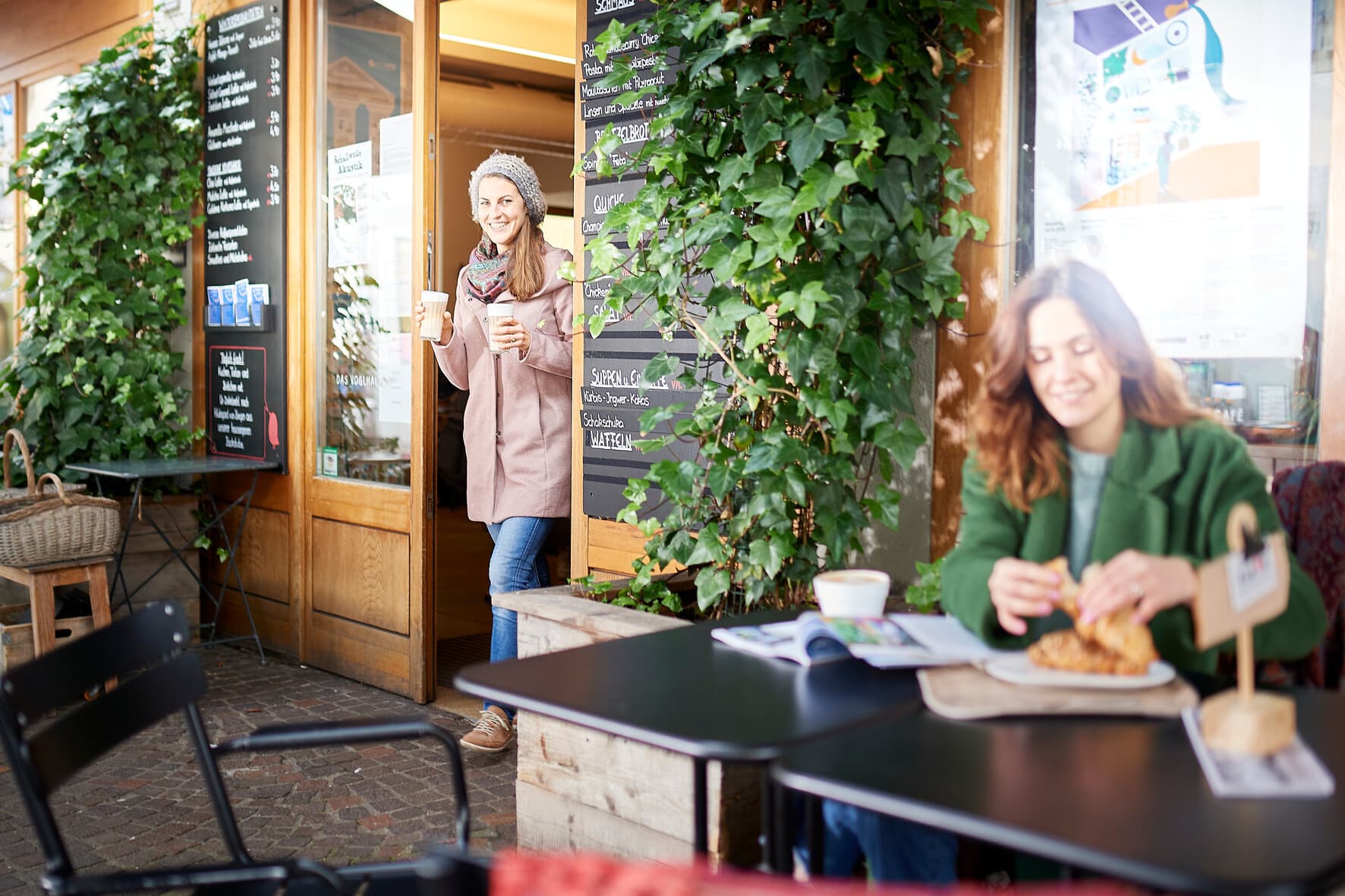 Blick auf die Tür des Voglhaus Cafés, aus der eine lächelnde Frau in Winterjacke mit 2 Kaffeebechern kommt.